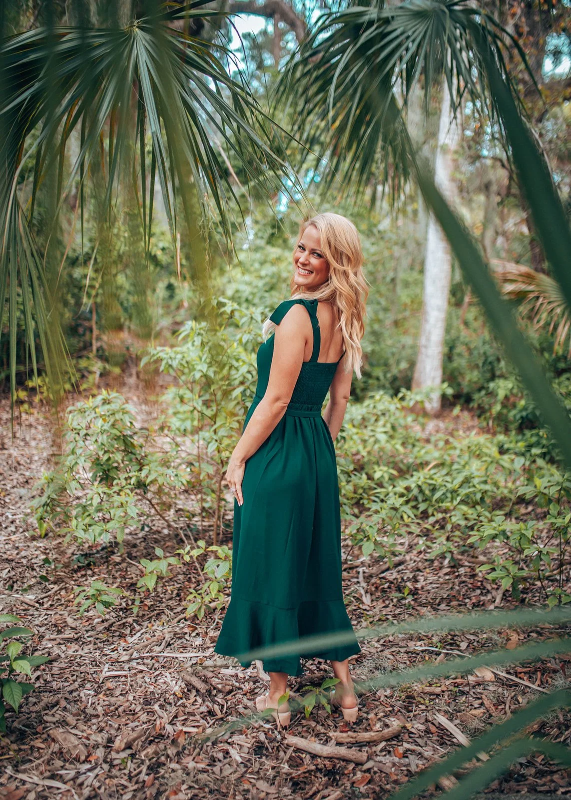 Sage Summer in green dress standing in a forest clearing surrounded by palm leaves.