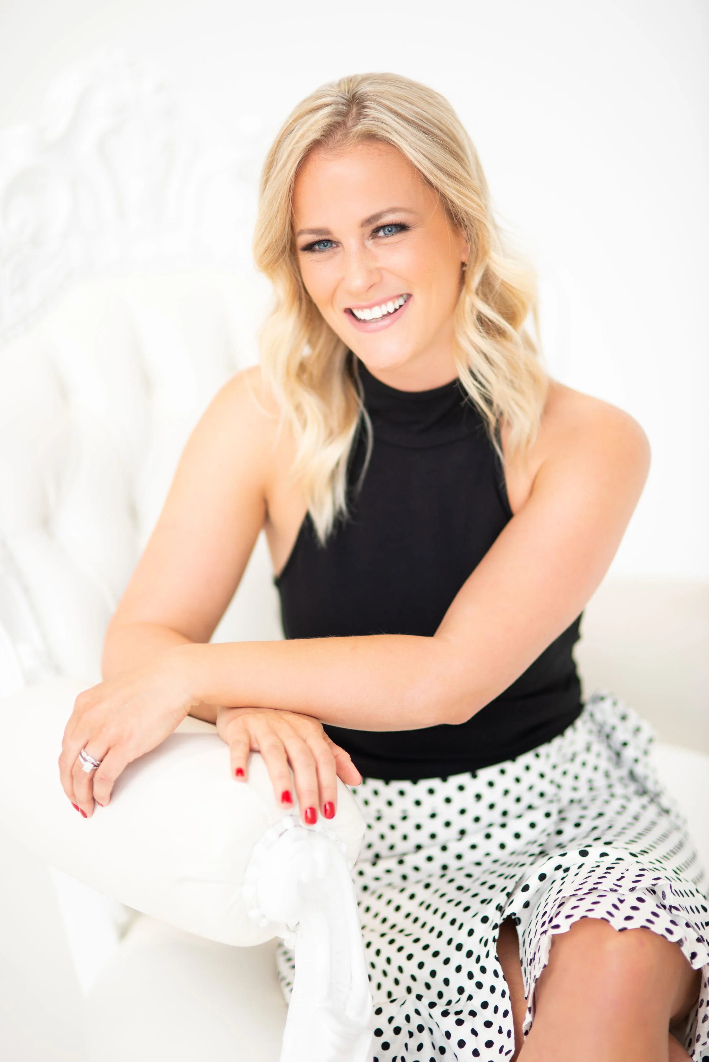 Sage Summer sitting on a white chair, wearing a black sleeveless top and a polka dot skirt, smiling.