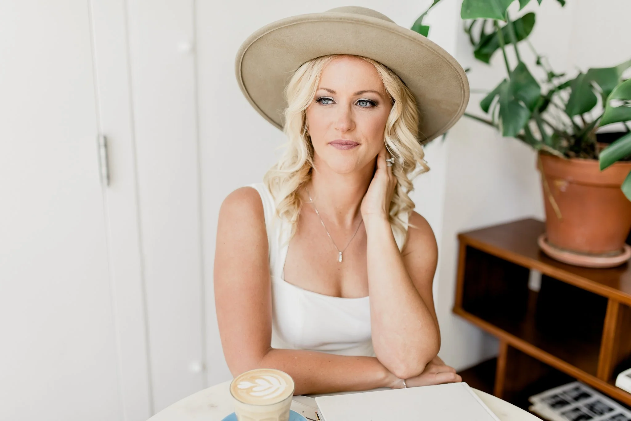 Sage Summer sitting at a table with a latte, wearing a wide-brimmed hat and white dress, with a plant in the background.