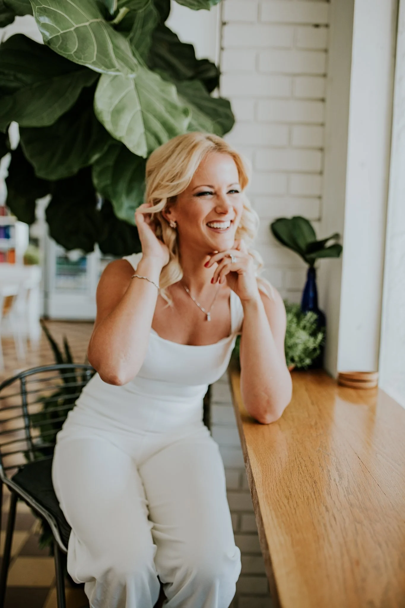 Sage Summer in white outfit smiling while sitting at a wooden counter, surrounded by plants.