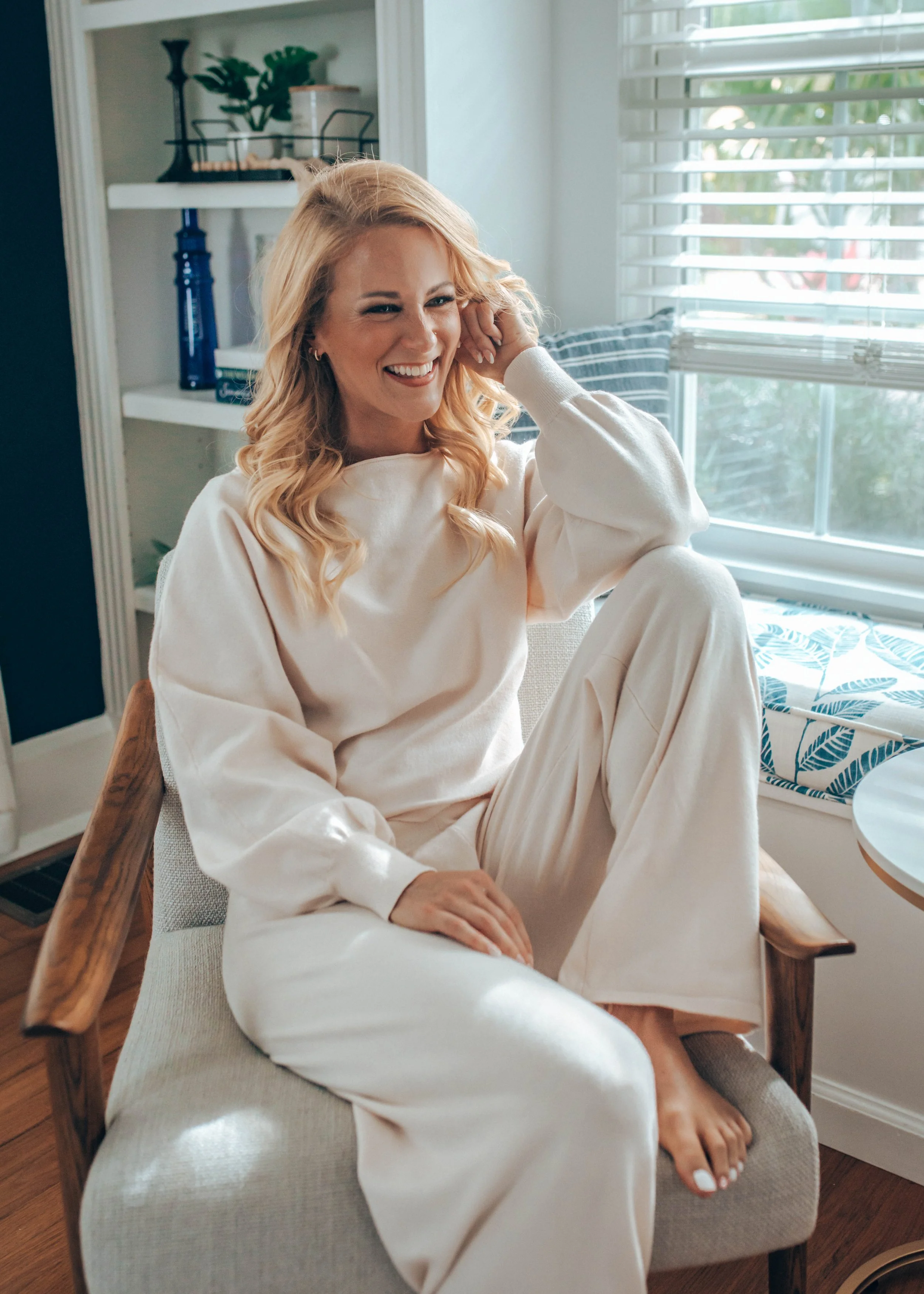 Sage Summer sitting on a chair, smiling and wearing a light-colored outfit in a sunlit room.