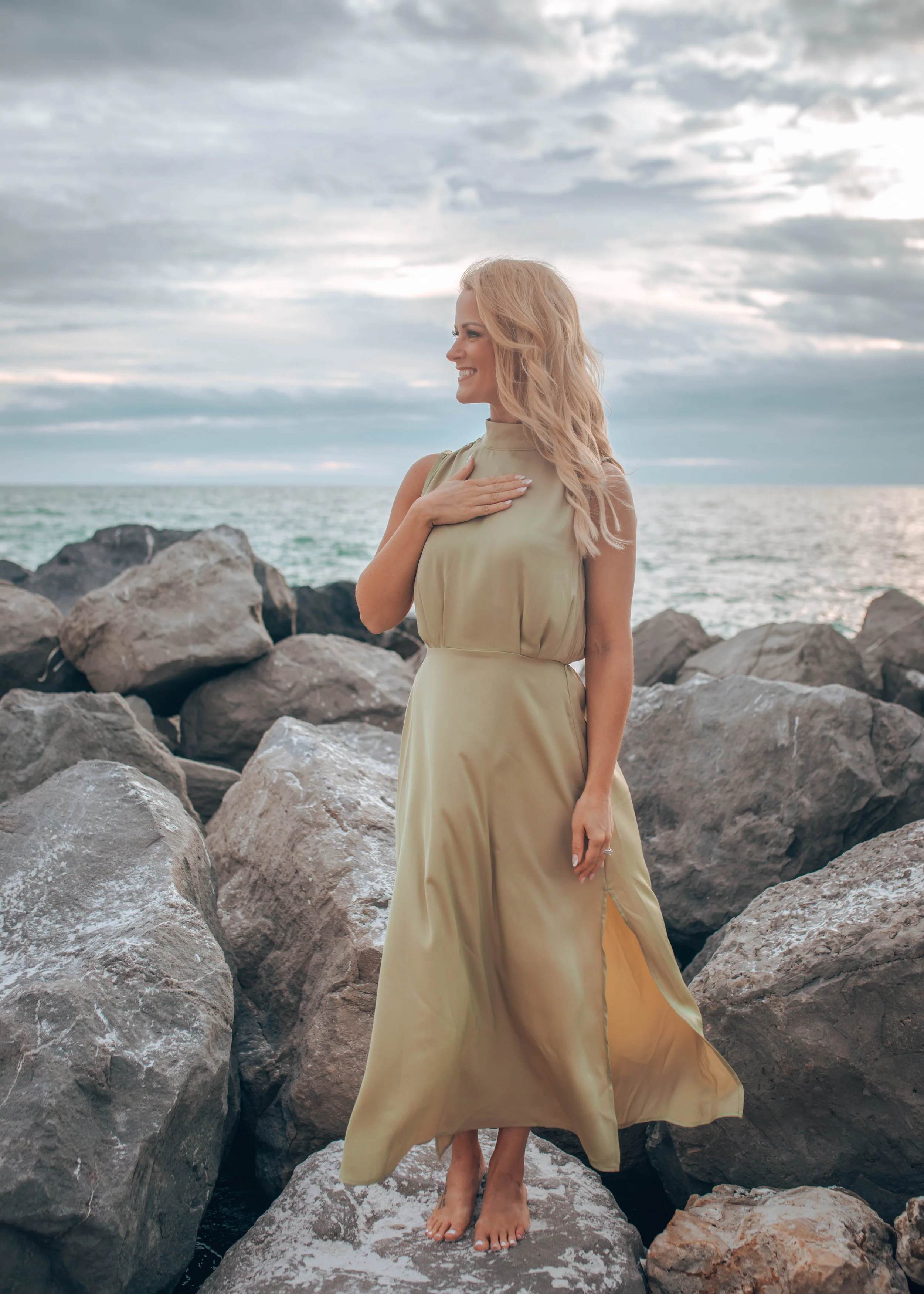 Sage Summer in green dress standing barefoot on rocks by the ocean, cloudy sky.