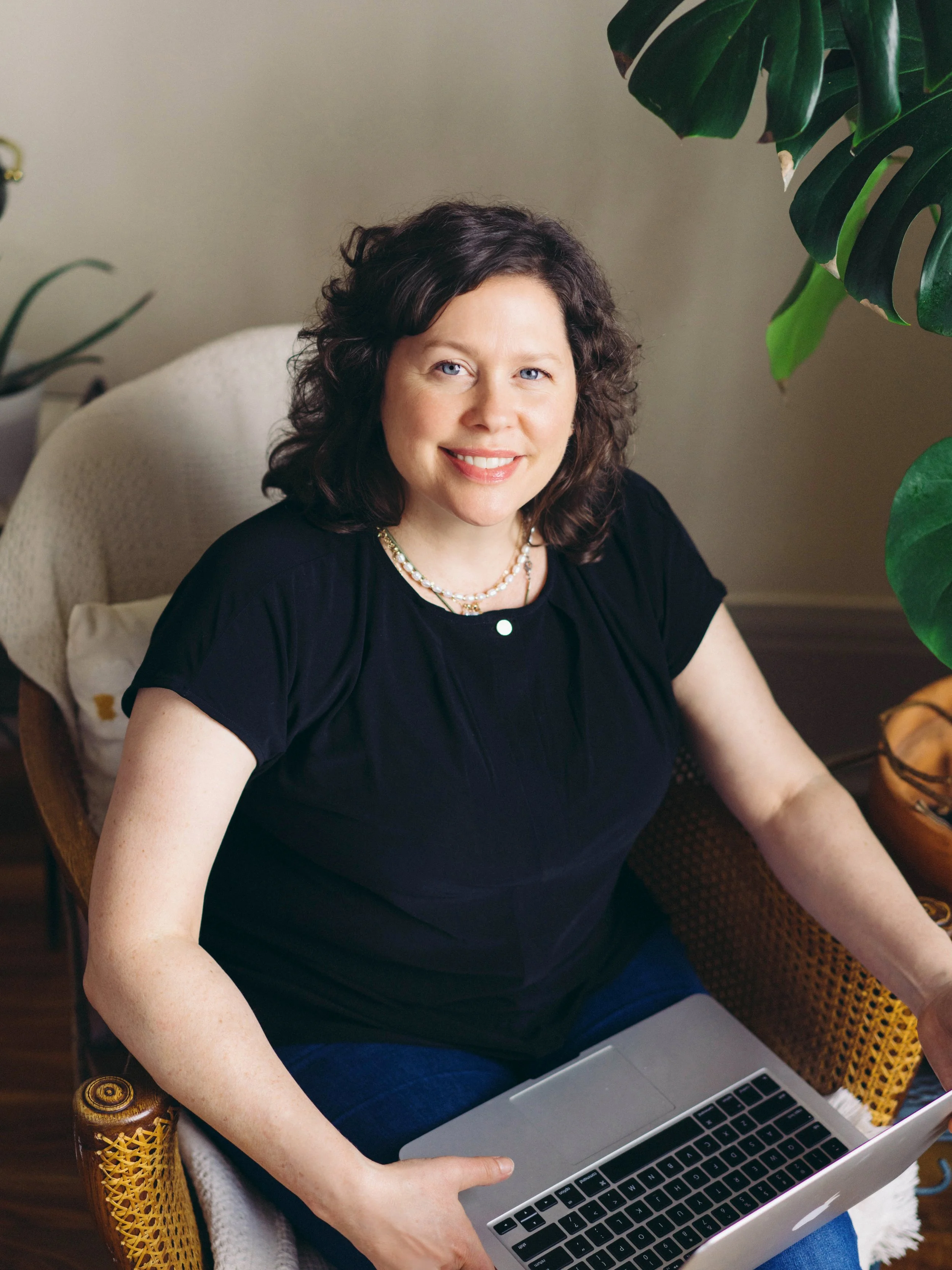 
shannon lu shan documentary & lifestyle family photographer Arnprior, Branding photography, A woman with curly dark hair and light skin sitting at a desk, smiling, and working on a laptop. 