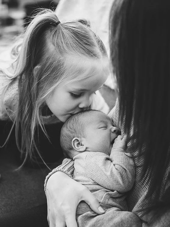 Shannonlushan Family newborn Photographer Arnprior - A young girl is kissing a sleeping baby's head while being held by an adult, with both in an outdoor setting.