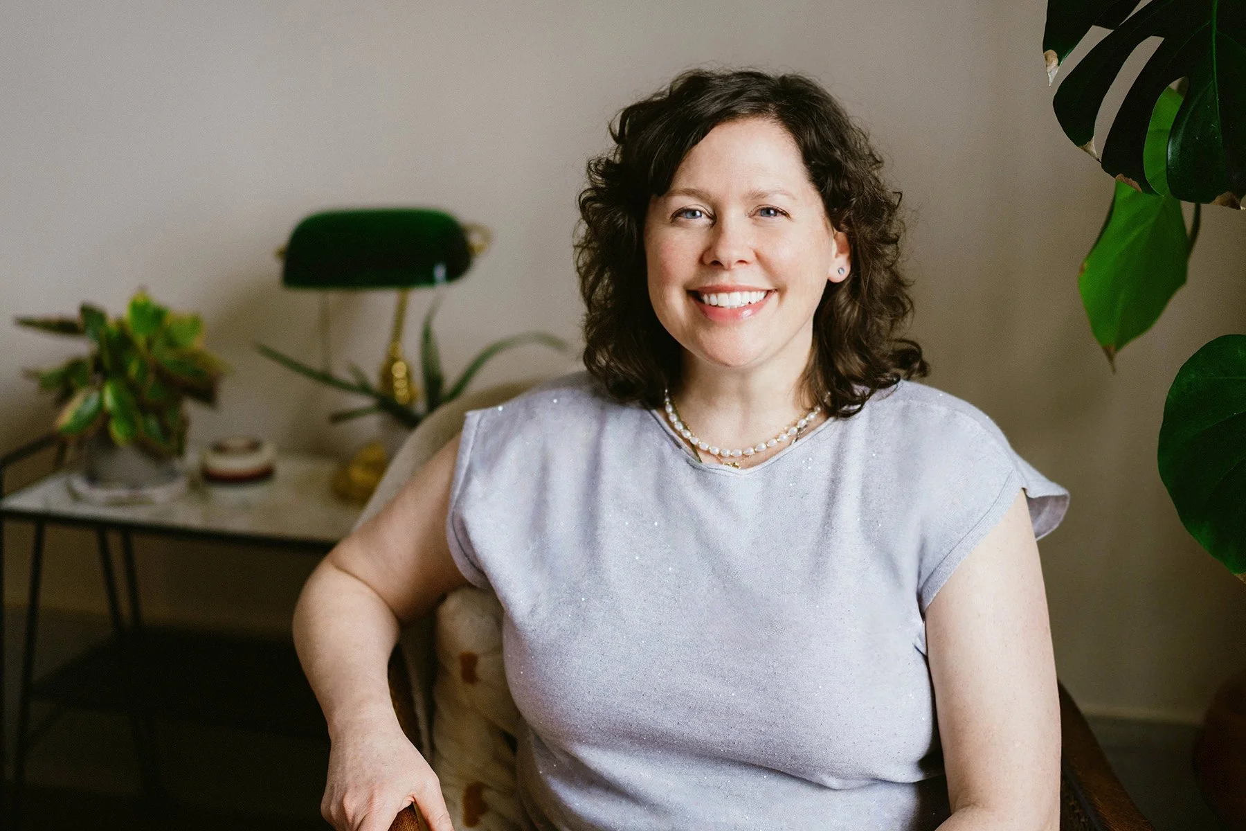 	
shannon lu shan documentary & lifestyle family photographer Arnprior , Branding photography A woman with shoulder-length curly brown hair smiling and sitting indoors, wearing a light purple sleeveless top and pearl necklace, 