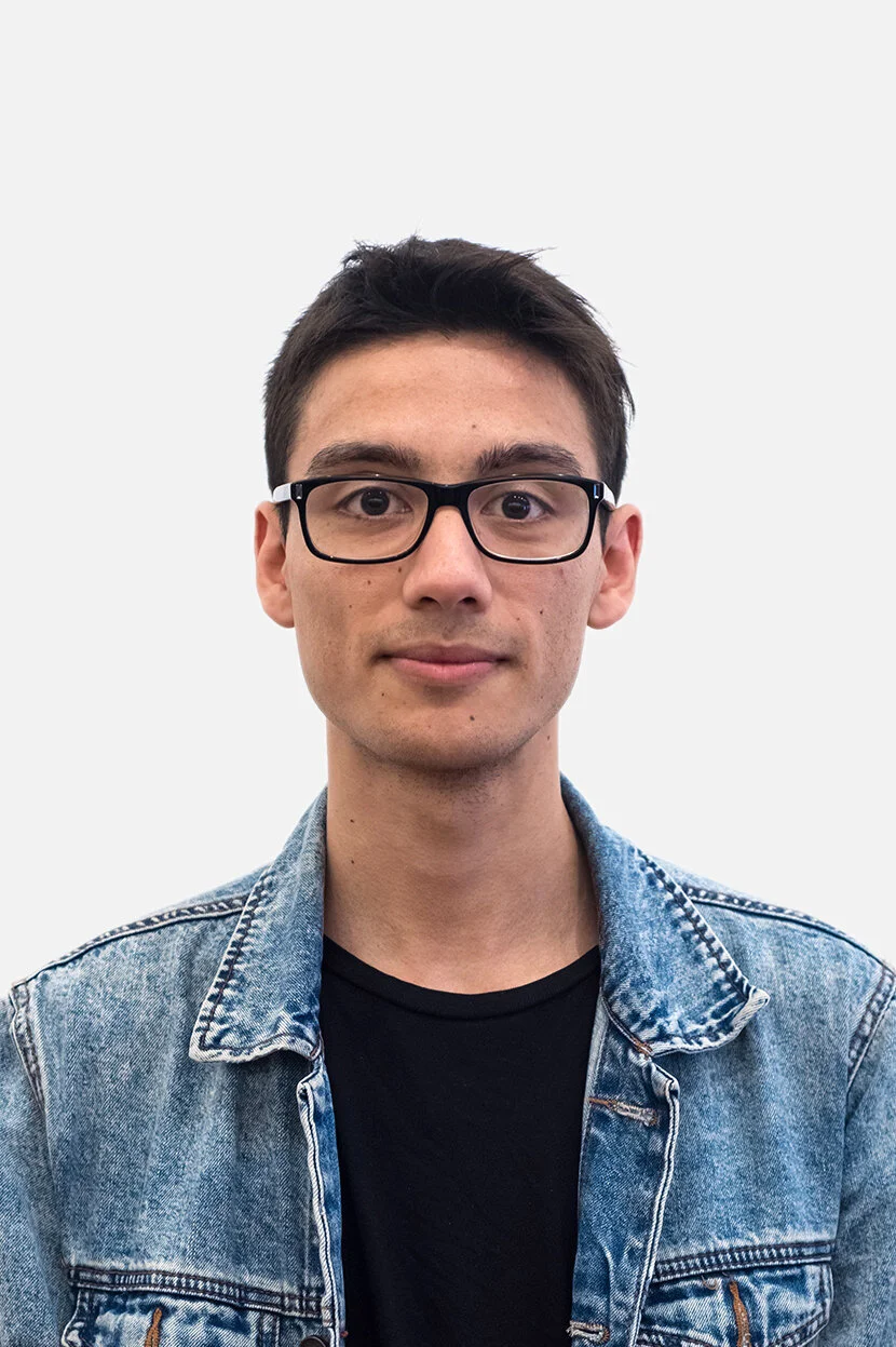 Portrait of a young man wearing glasses and a denim jacket against a plain light background.