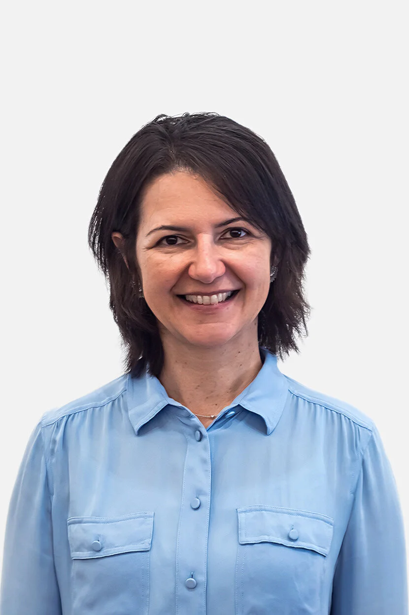 A woman with dark brown hair in a blue button-up shirt, smiling, against a plain white background.