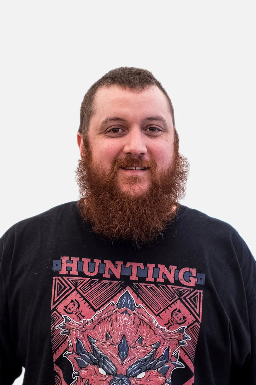 A man with a beard and short hair wearing a black T-shirt with a graphic and the word 'HUNTING' on it, smiling and standing against a plain light background.