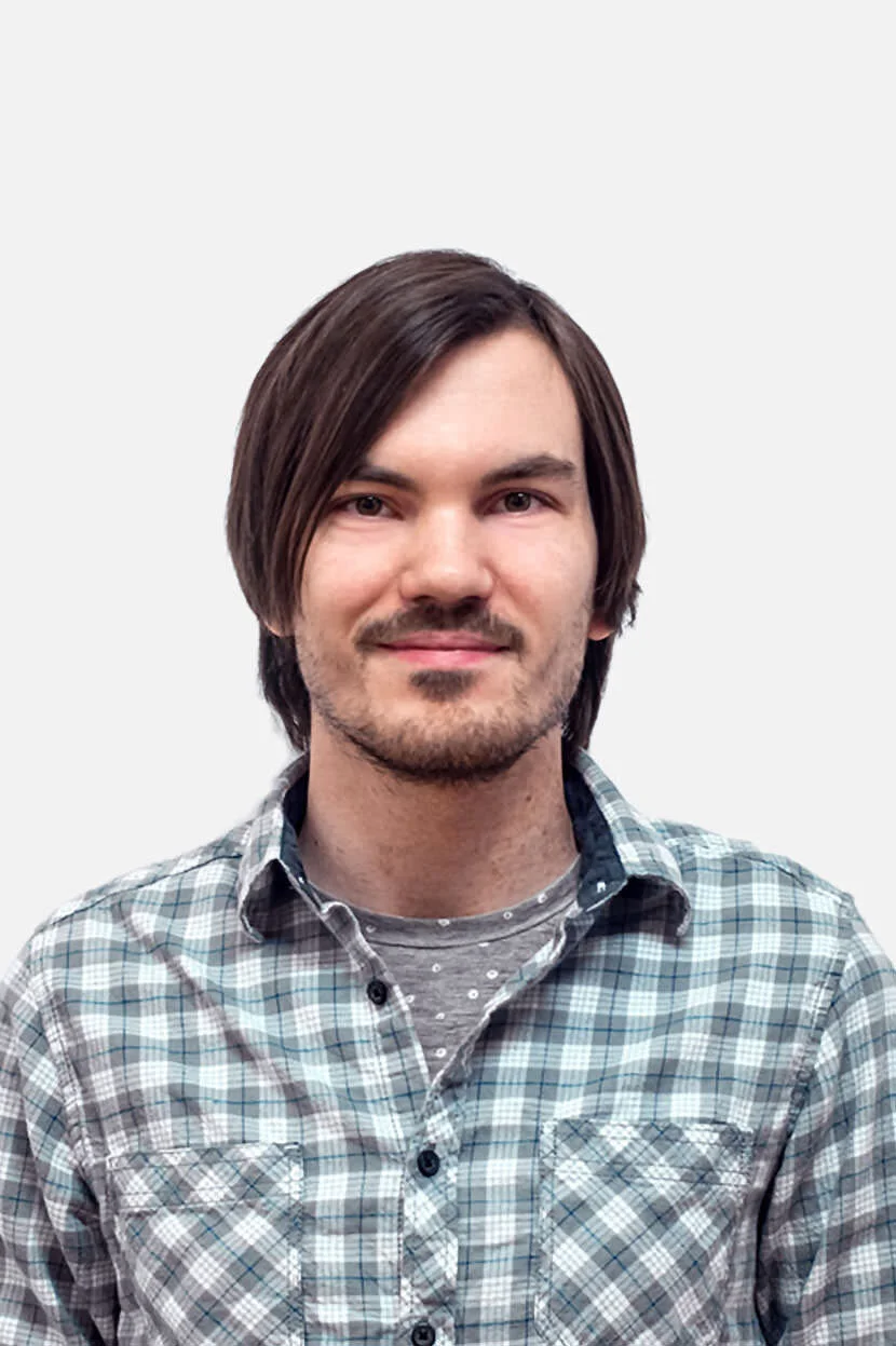 Portrait of a young man with brown hair, light stubble, wearing a gray plaid shirt over a gray t-shirt with white polka dots, against a plain white background.