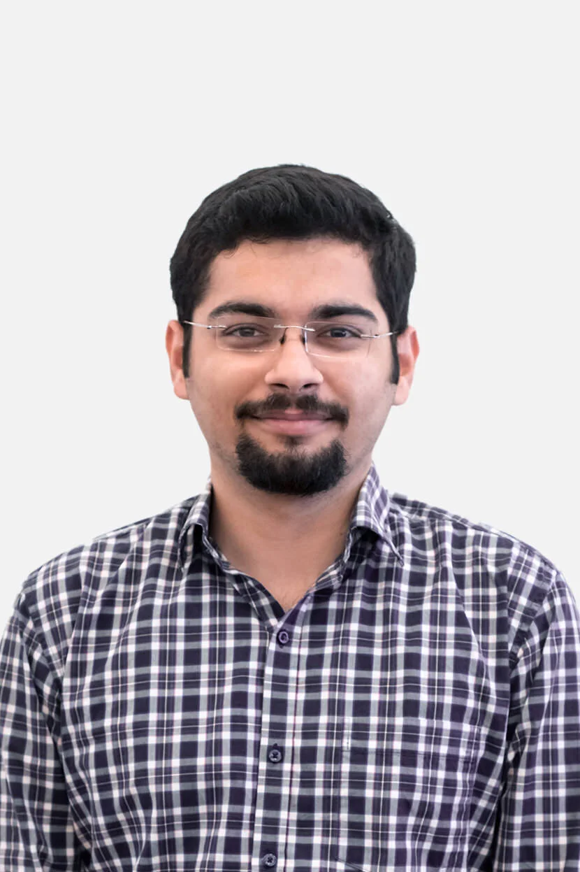 Portrait of a young man with glasses, short dark hair, a goatee, smiling, wearing a checkered shirt, against a plain white background.