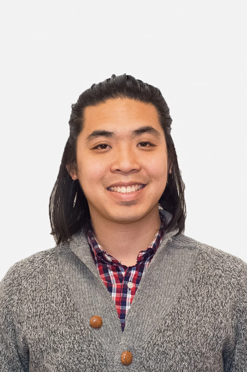 Young man with shoulder-length black hair, smiling, wearing a gray knit sweater and a red, blue, and white plaid shirt against a white background.