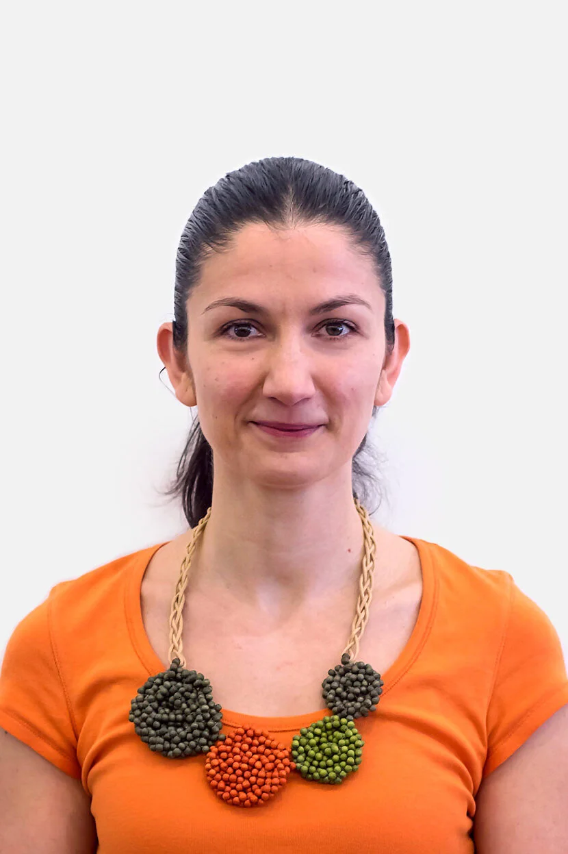 A woman wearing an orange shirt and a chunky necklace with large clustered beads in black, orange, and green, standing in front of a plain white background.