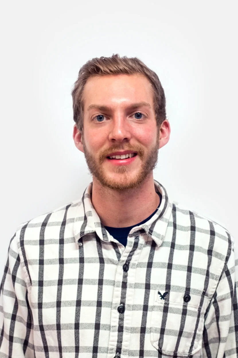 A young man with light skin, light brown hair, and a beard, smiling, wearing a black and white plaid shirt.