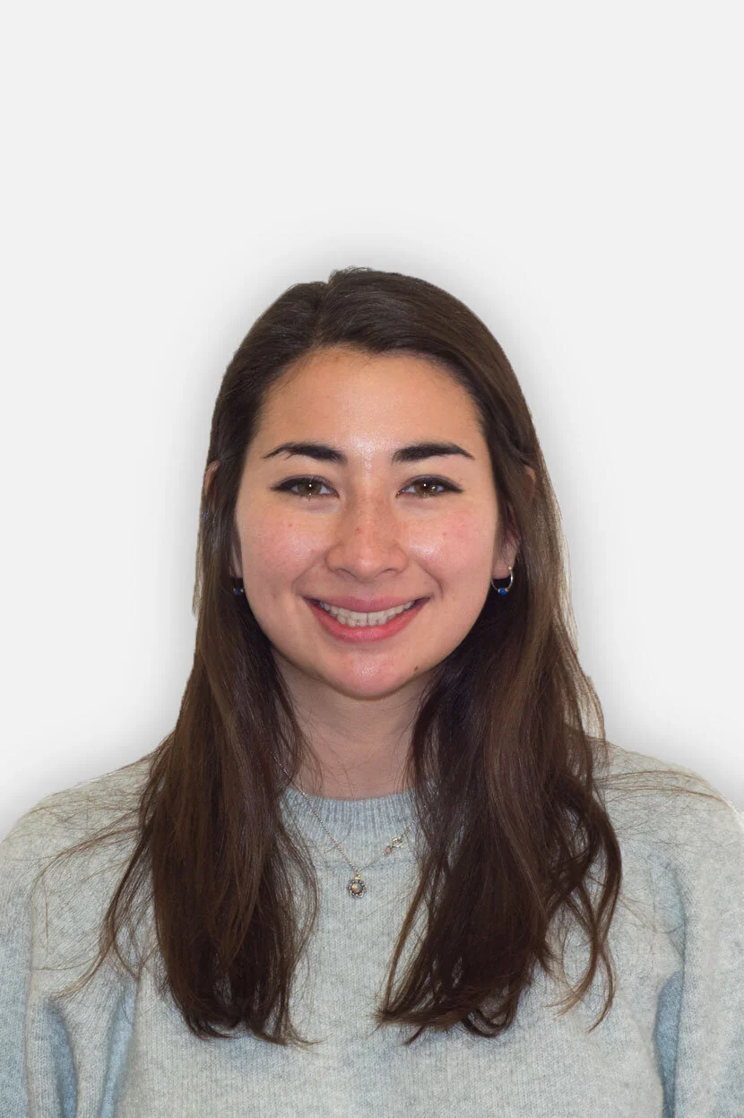A young woman with long brown hair, smiling, wearing a light grey sweater and jewelry, against a plain light background.