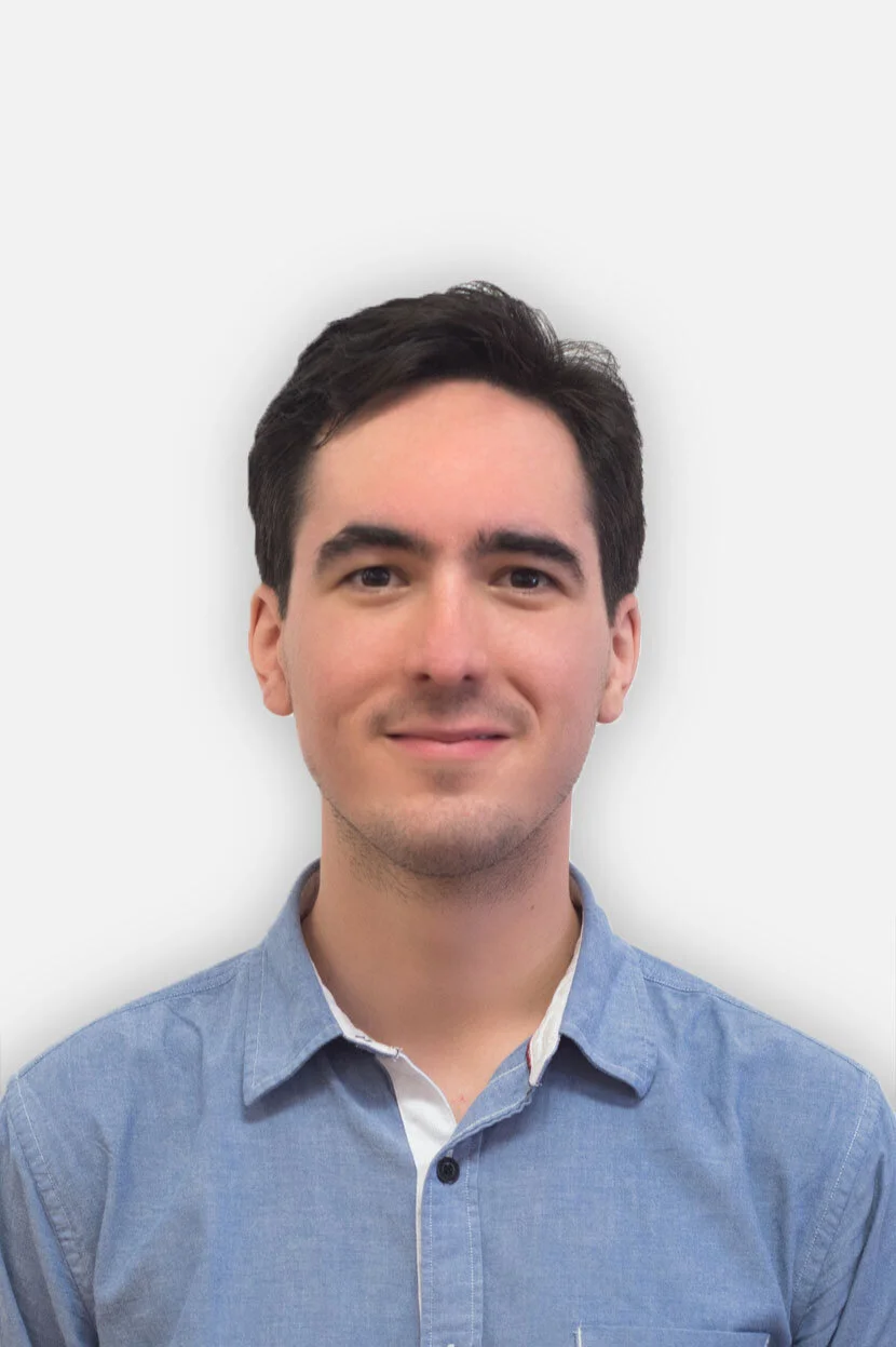 Portrait of a young man with dark hair wearing a light blue button-up shirt against a plain white background.