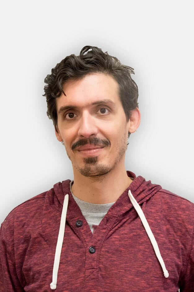 A young man with wavy dark hair, facial hair, and light skin, wearing a maroon hoodie with white drawstrings and a gray T-shirt, standing against a light background.