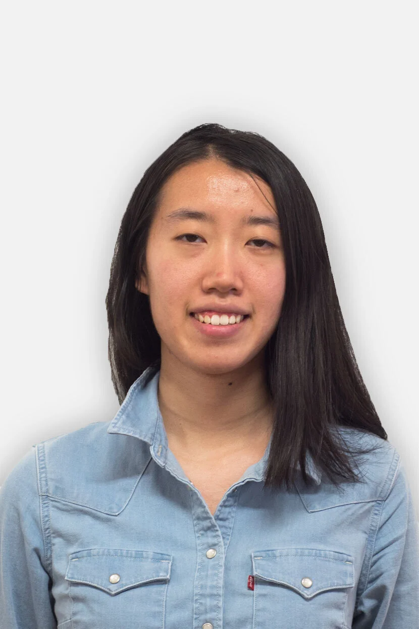 A young woman with shoulder-length black hair wearing a light blue denim shirt, smiling in front of a plain white background.