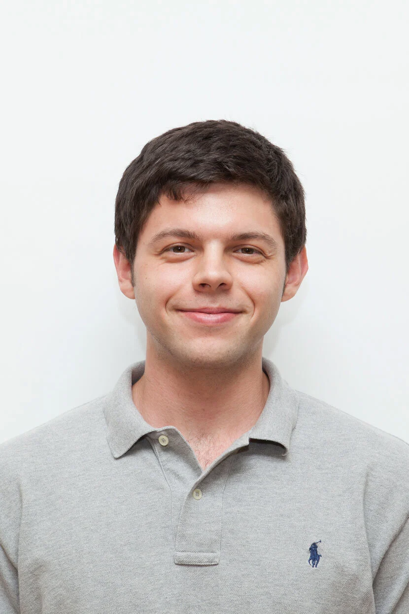 A young man with dark hair smiling, wearing a light gray polo shirt with a small polo logo, standing against a plain white background.