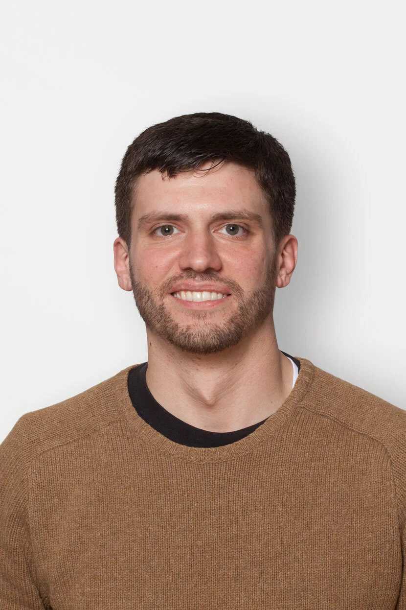 Portrait of a young man with short dark hair, a beard, and blue eyes, smiling, wearing a tan sweater with a black shirt underneath, standing against a plain white background.
