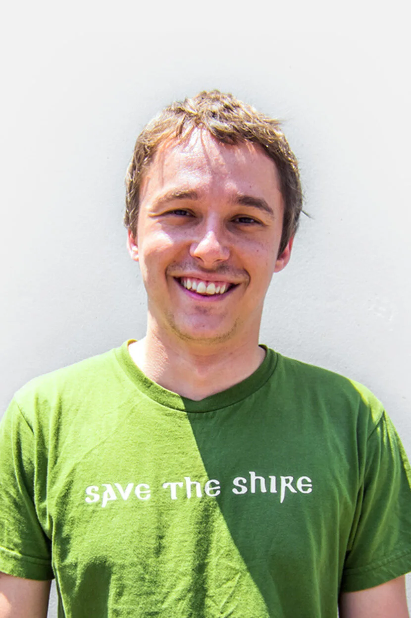 A young man smiling outdoors wearing a green T-shirt that says 'SAVE THE SHIRE'.
