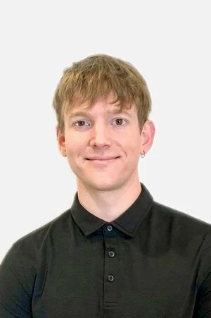 A young man with light brown hair, wearing a black collared shirt, smiling against a plain white background.