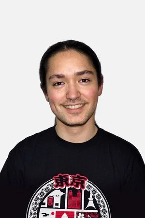 A young man with dark hair and a slight beard smiling, wearing a black T-shirt with a colorful graphic design.