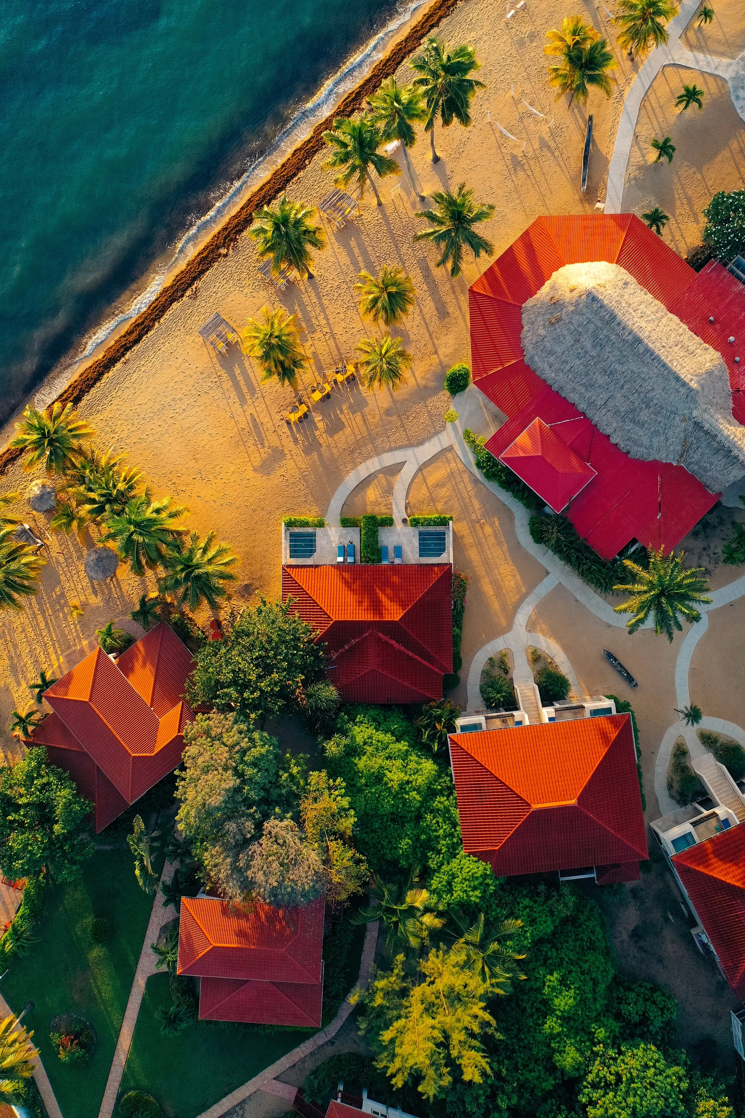 Aerial view of a beach resort with red-roofed buildings, palm trees, and a sandy shoreline along a body of water.