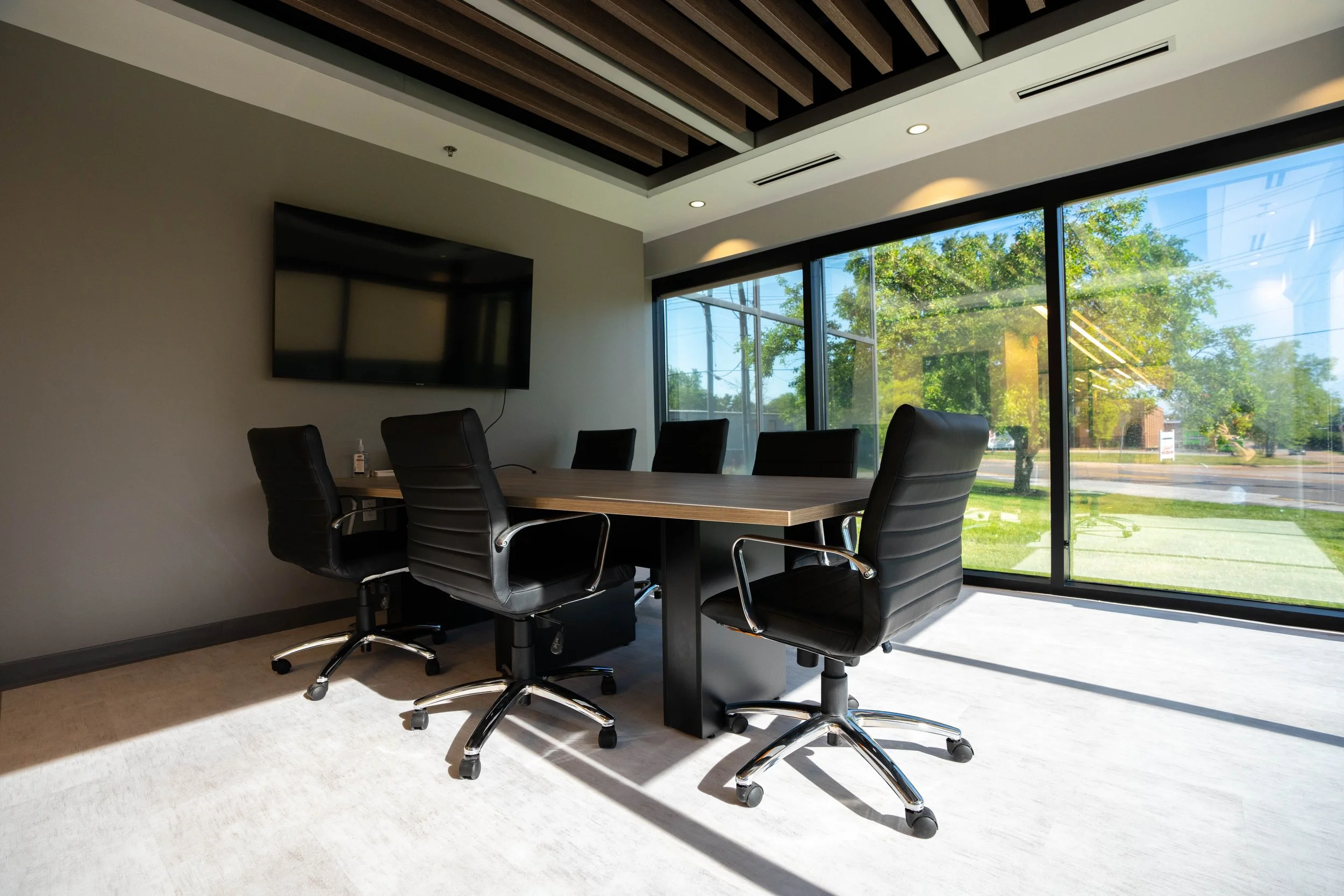 Modern conference room with a long table, five black leather chairs, large windows with a view of trees, and a flat-screen TV mounted on the wall.