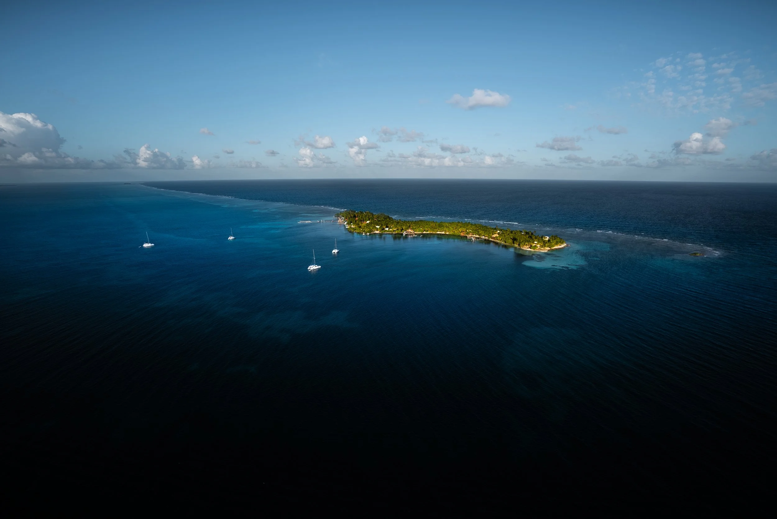 Aerial view of a small tropical island surrounded by blue ocean with several sailboats nearby under a clear sky.