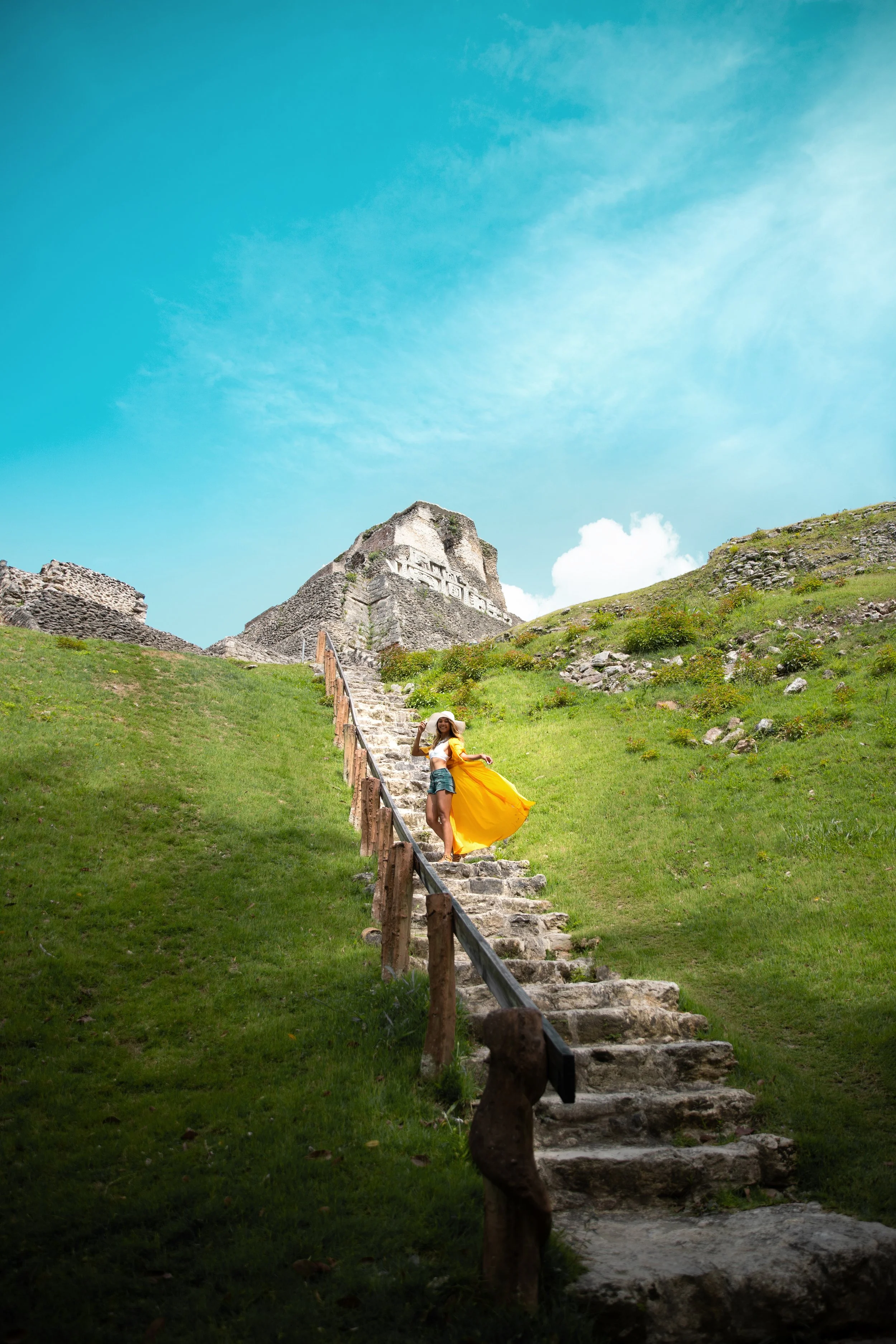 A stone pathway leading up a grassy hill toward ancient ruins, with a person in a bright yellow dress and hat standing on the steps.