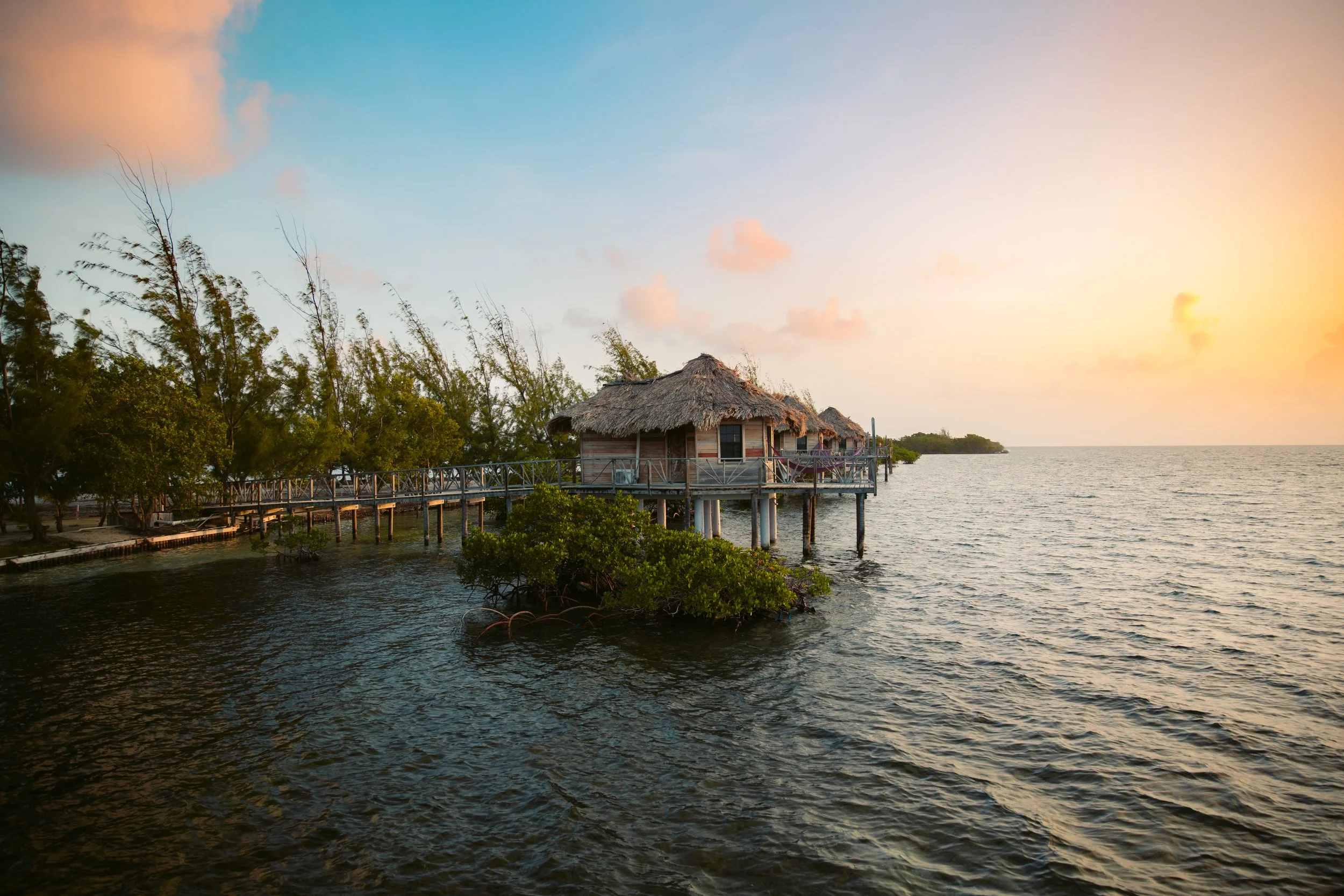Thatched-roof huts on stilts over water at sunset, with a wooden walkway and surrounding trees.
