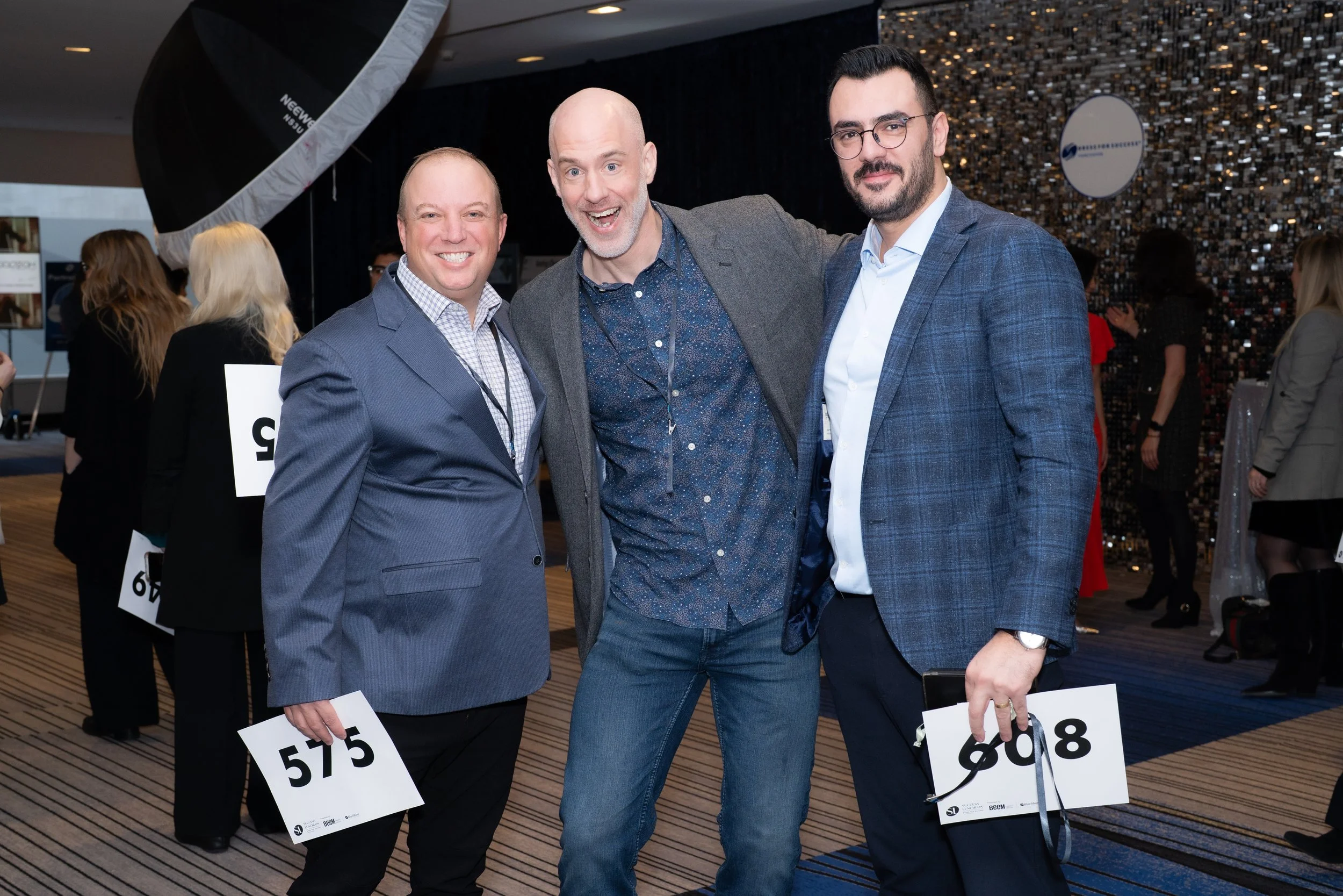 Three men standing together at an event, smiling at the camera. The man on the left is wearing a blue suit with a checkered shirt, the man in the middle is bald with a gray beard, wearing a gray jacket over a patterned blue shirt, and the man on the 