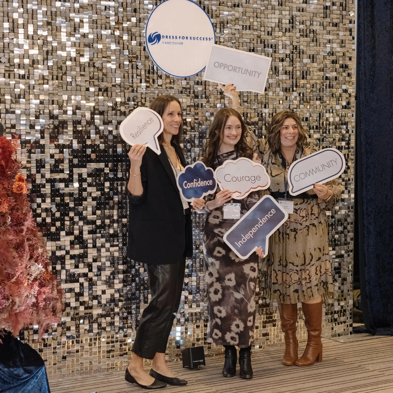 Three women standing in front of a silver mosaic wall, holding signs with words like 'Resilience,' 'Confidence,' 'Courage,' 'Independence,' 'Community,' and 'Opportunity.' The event is organized by 'Dress for Success Vancouver.'