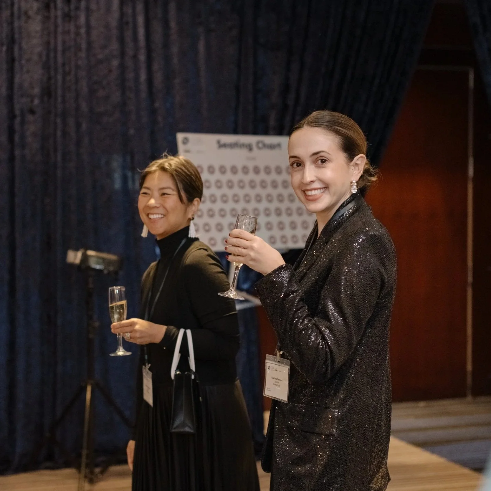 Two women smiling and holding champagne glasses at an indoor event, with dark curtains and a sign in the background.