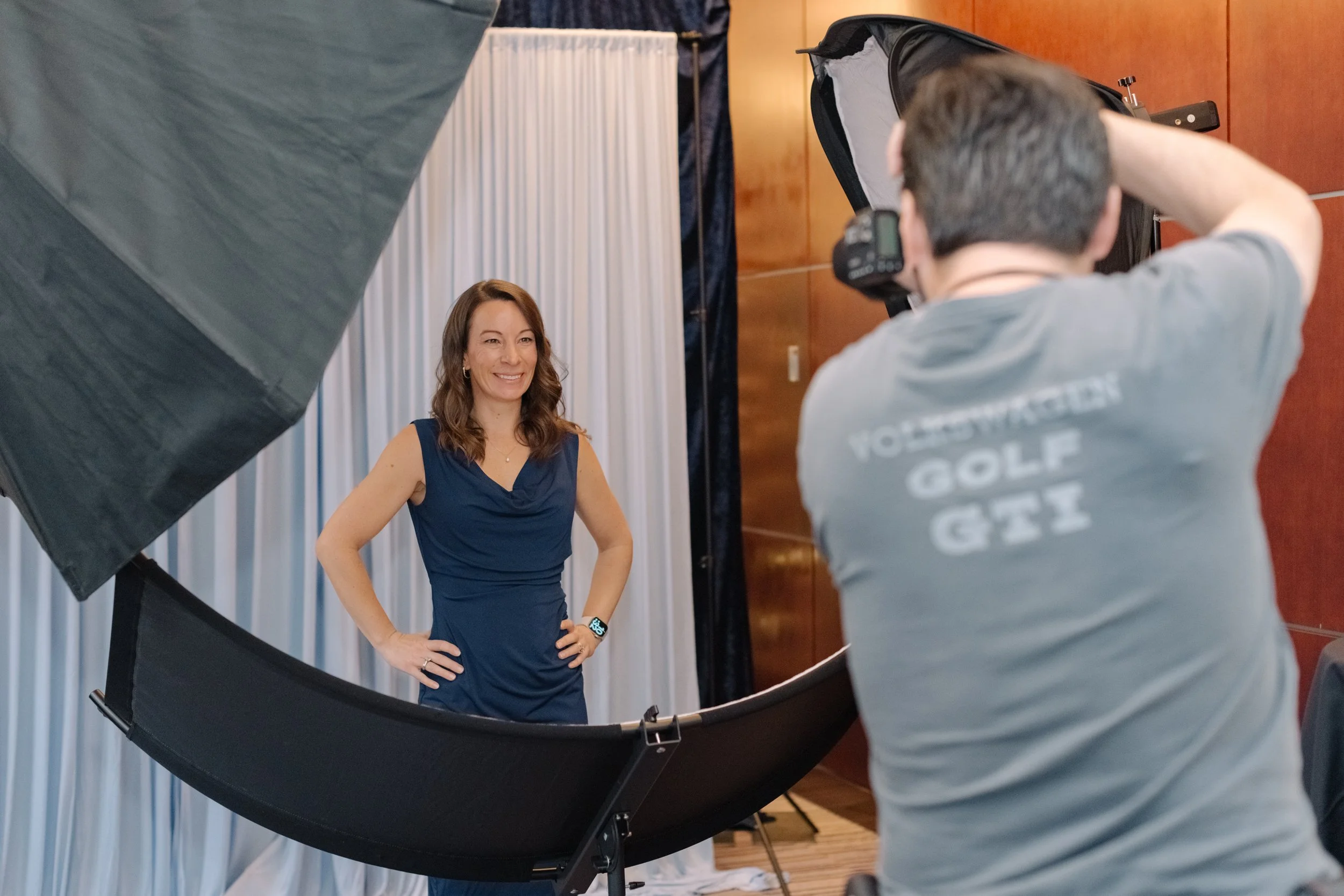 Woman in a navy blue dress smiling in a photography studio, being photographed by a man in a grey T-shirt behind a camera.