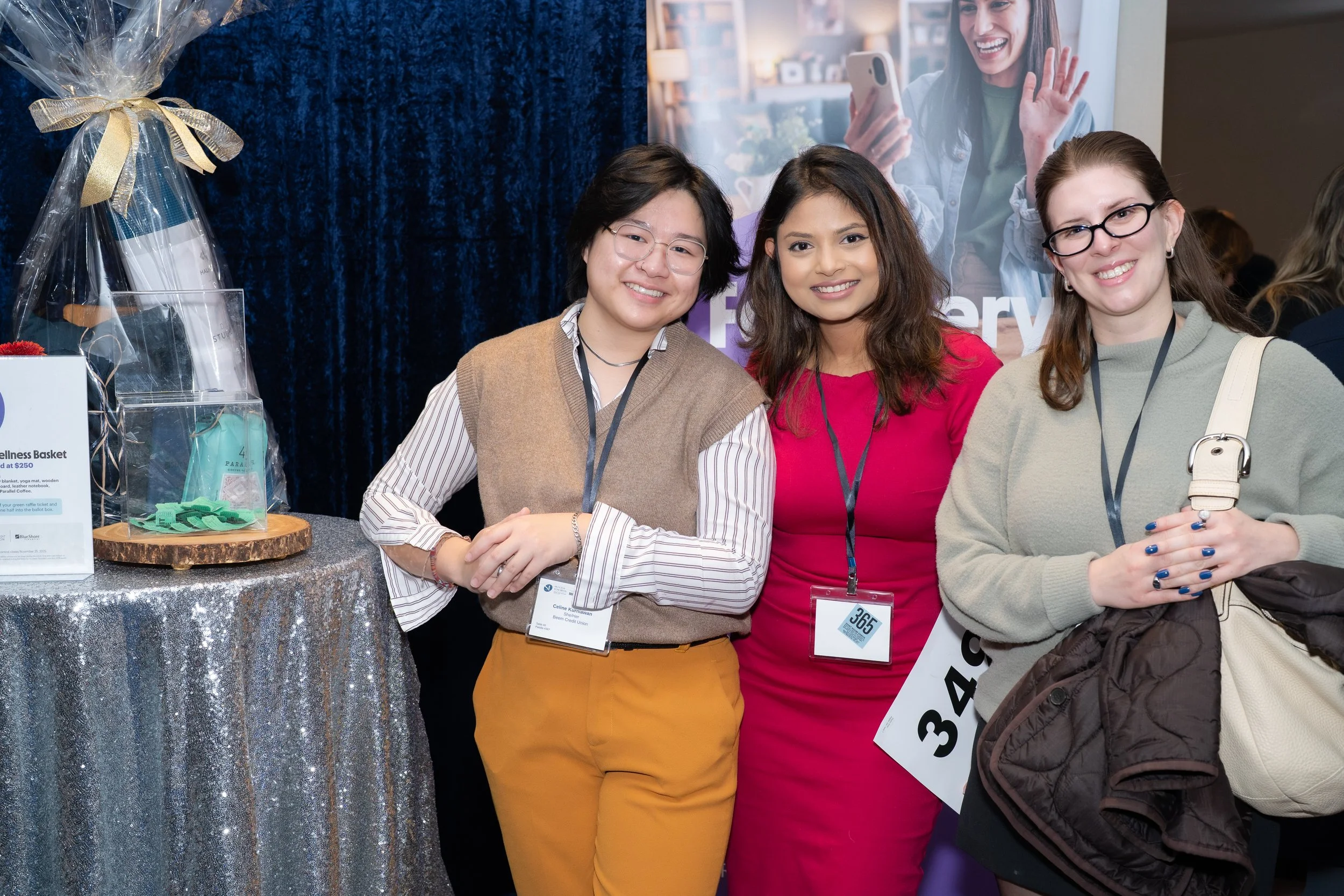 Three women standing together at a conference, smiling at the camera, wearing conference badges. One woman is in a beige vest and yellow pants, the second woman in a pink dress, and the third woman in a gray sweater holding a jacket and backpack.