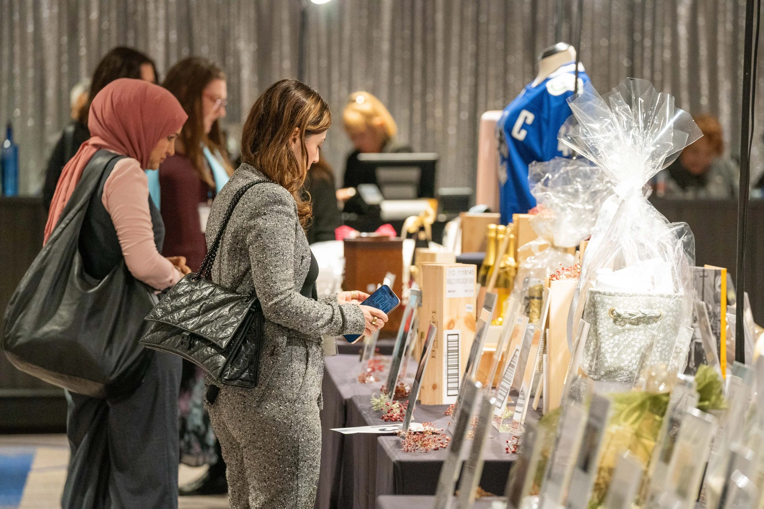 People browsing auction items at a charity event, with a woman in a gray suit in the foreground using her phone.