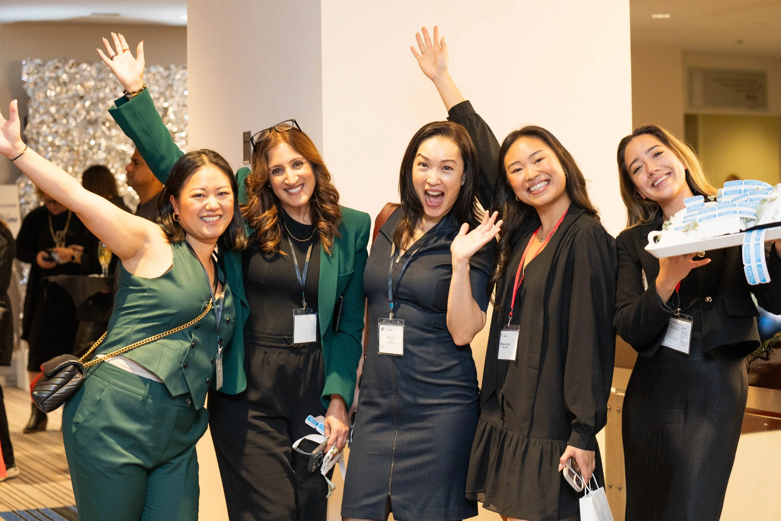 Group of five women smiling, celebrating, and waving at an event, wearing business attire and name badges.
