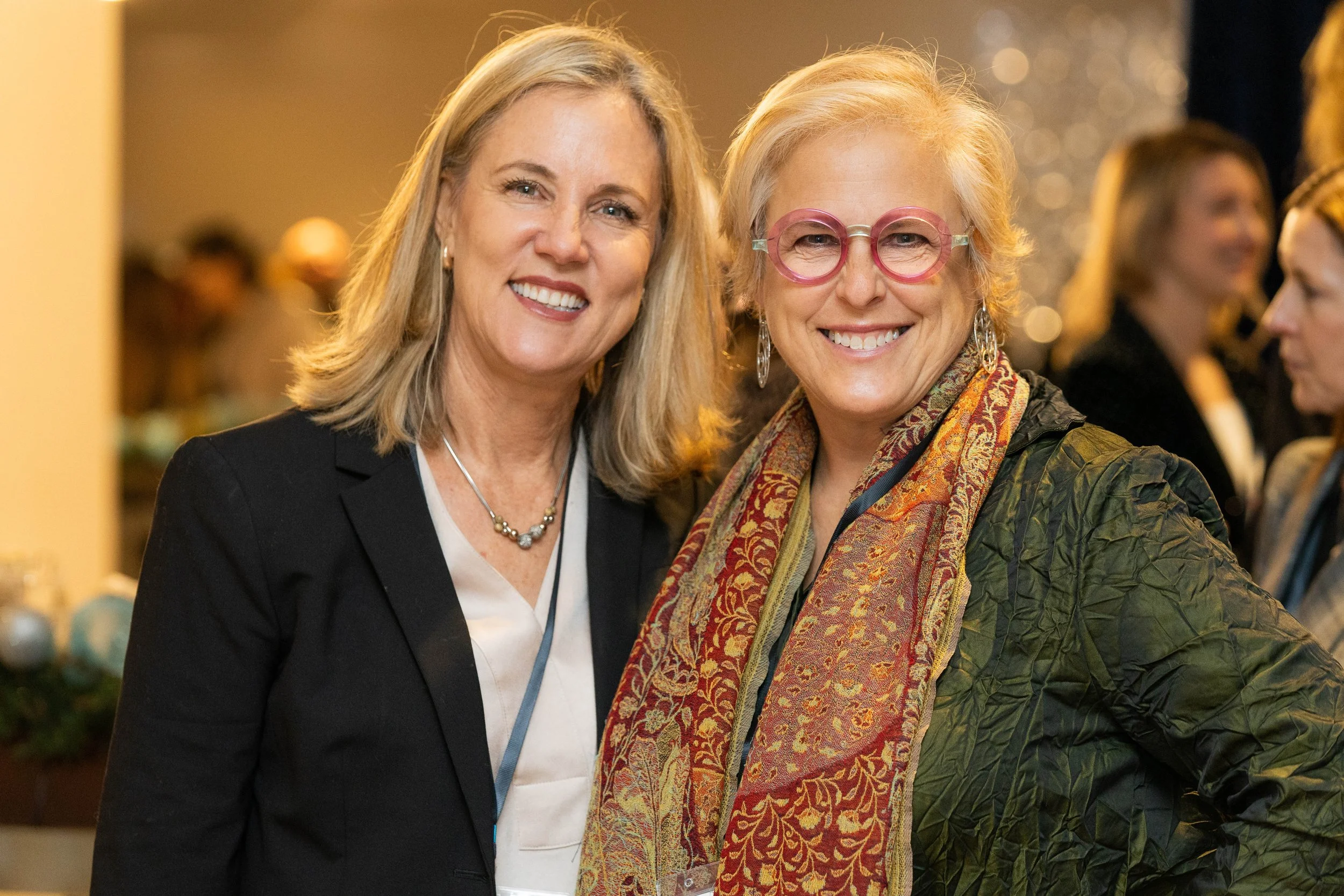 Two women smiling at an indoor event, one with blonde hair wearing a black blazer and pearls, and the other with curly blonde hair, pink glasses, and colorful scarf, standing close together.