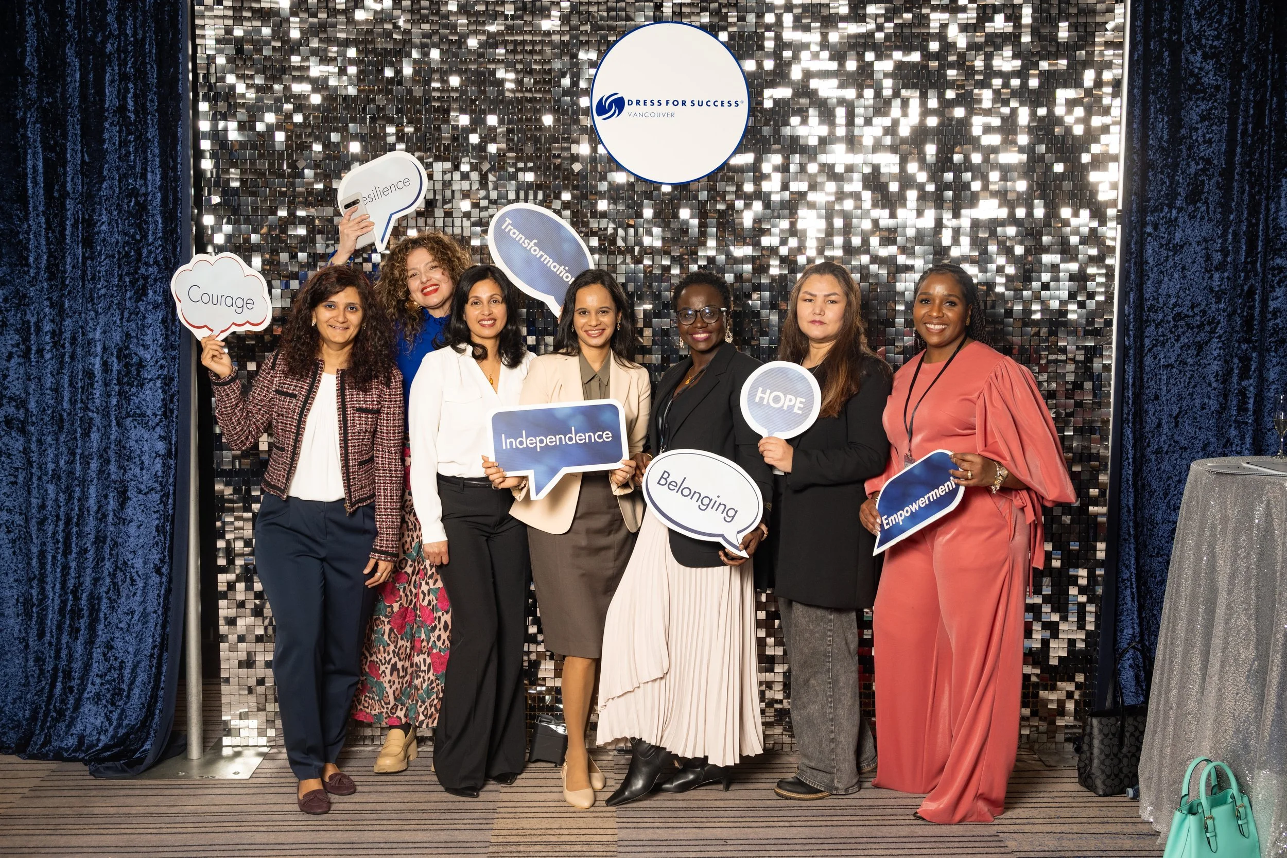 Group of women at a professional event holding speech bubbles with words like "Courage," "Resilience," "Independence," "Belonging," "Hope," and "Empowerment," standing in front of a sparkly black background with a round sign that reads "Dress for Success Vancouver."