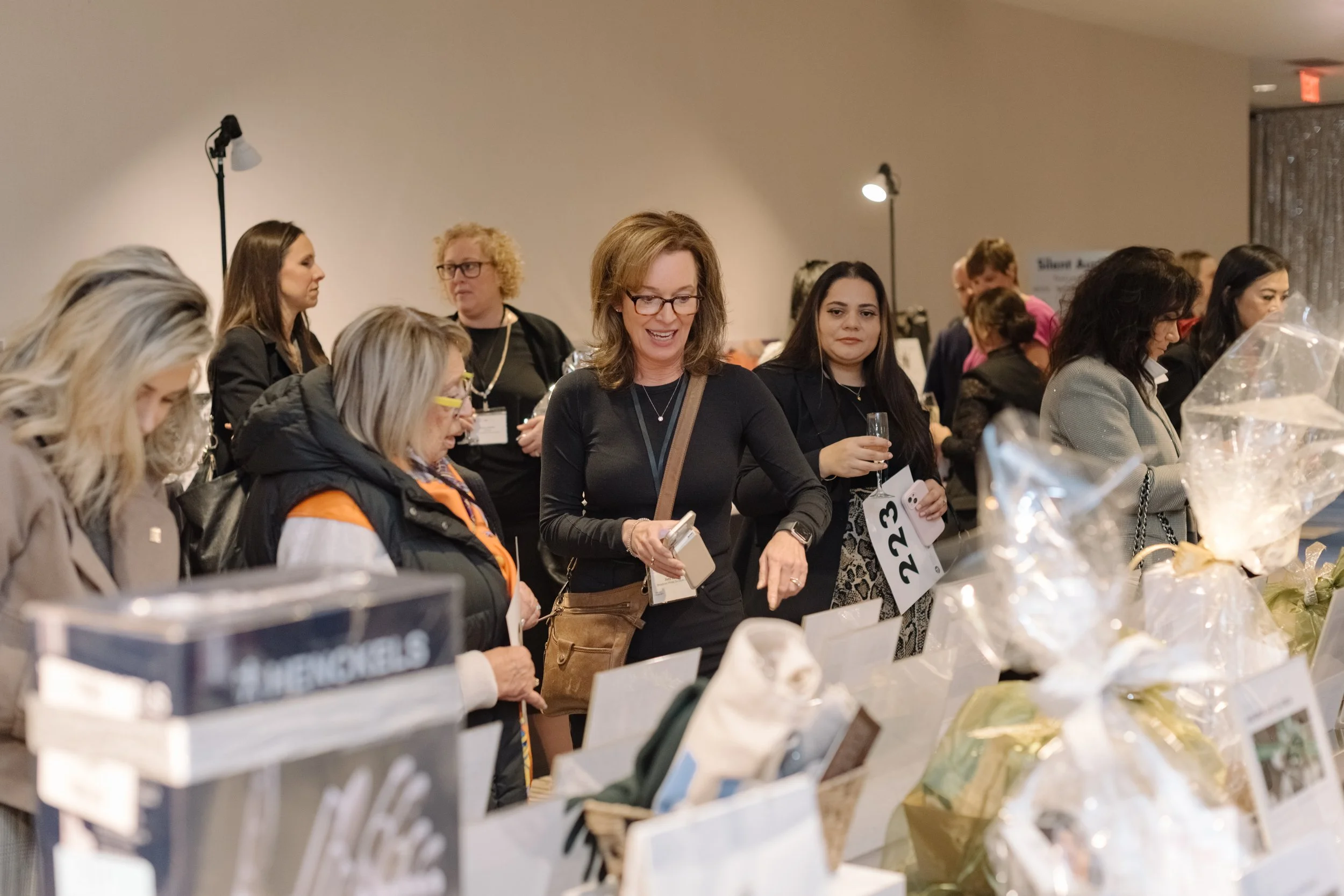 People at an event browsing items on a table, some holding drinks and reading printed materials, with a lively atmosphere.