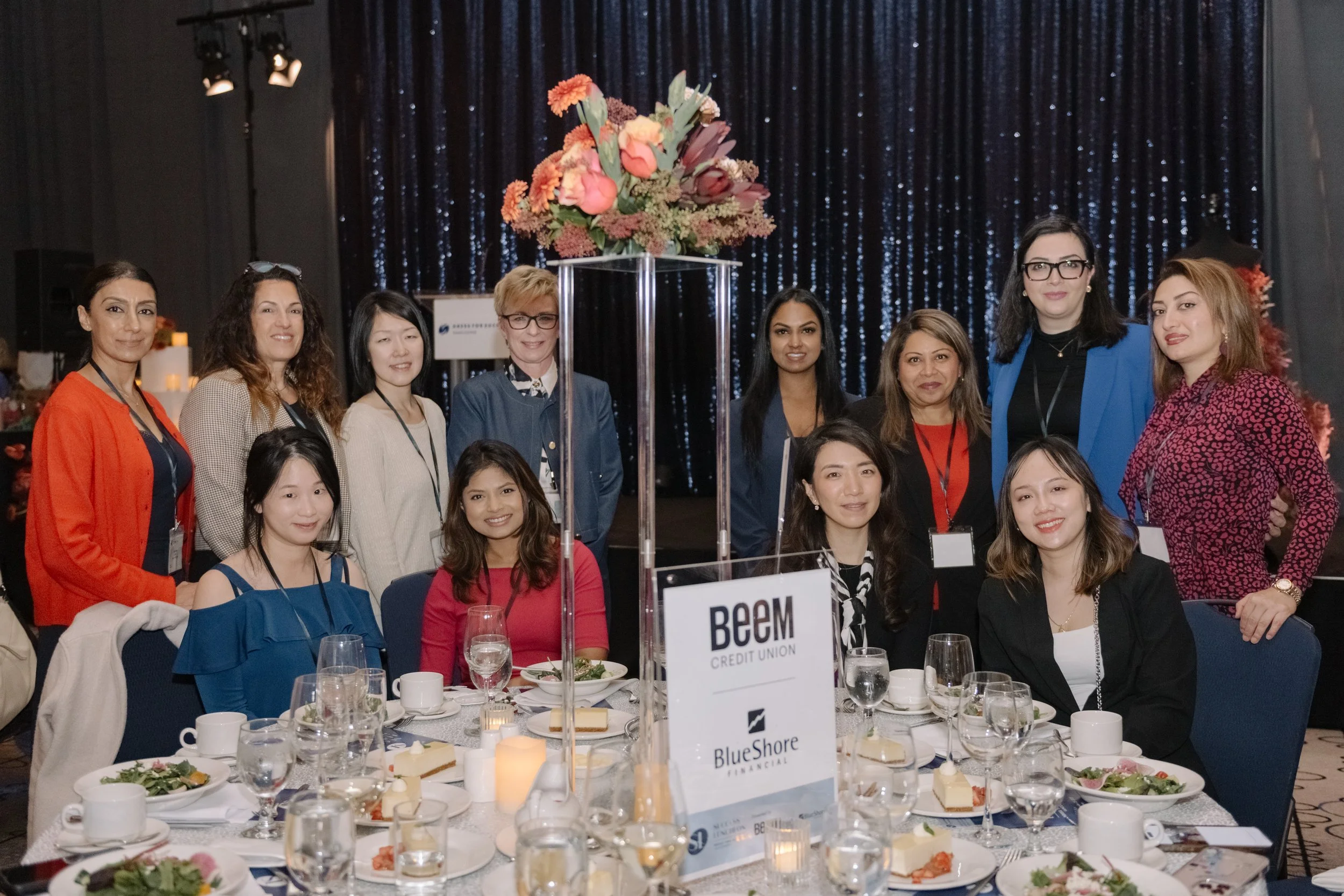 Group of women gathered at a formal event, seated around a table with food and drinks, with a tall floral centerpiece, in front of a dark, sparkly curtain background.