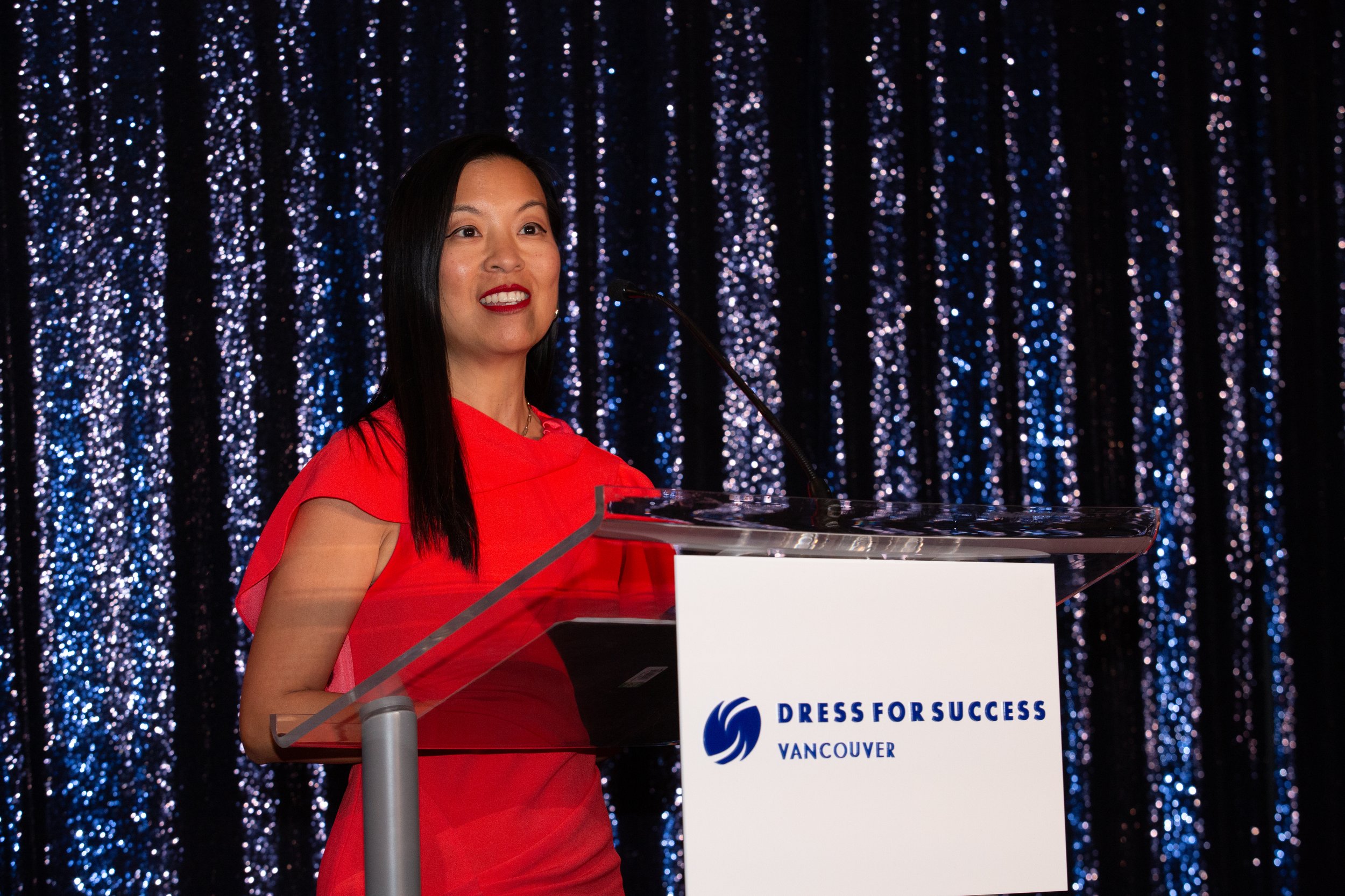A woman in a red dress speaking at a podium with a sign that reads 'Dress for Success Vancouver' and a sparkly black curtain background.