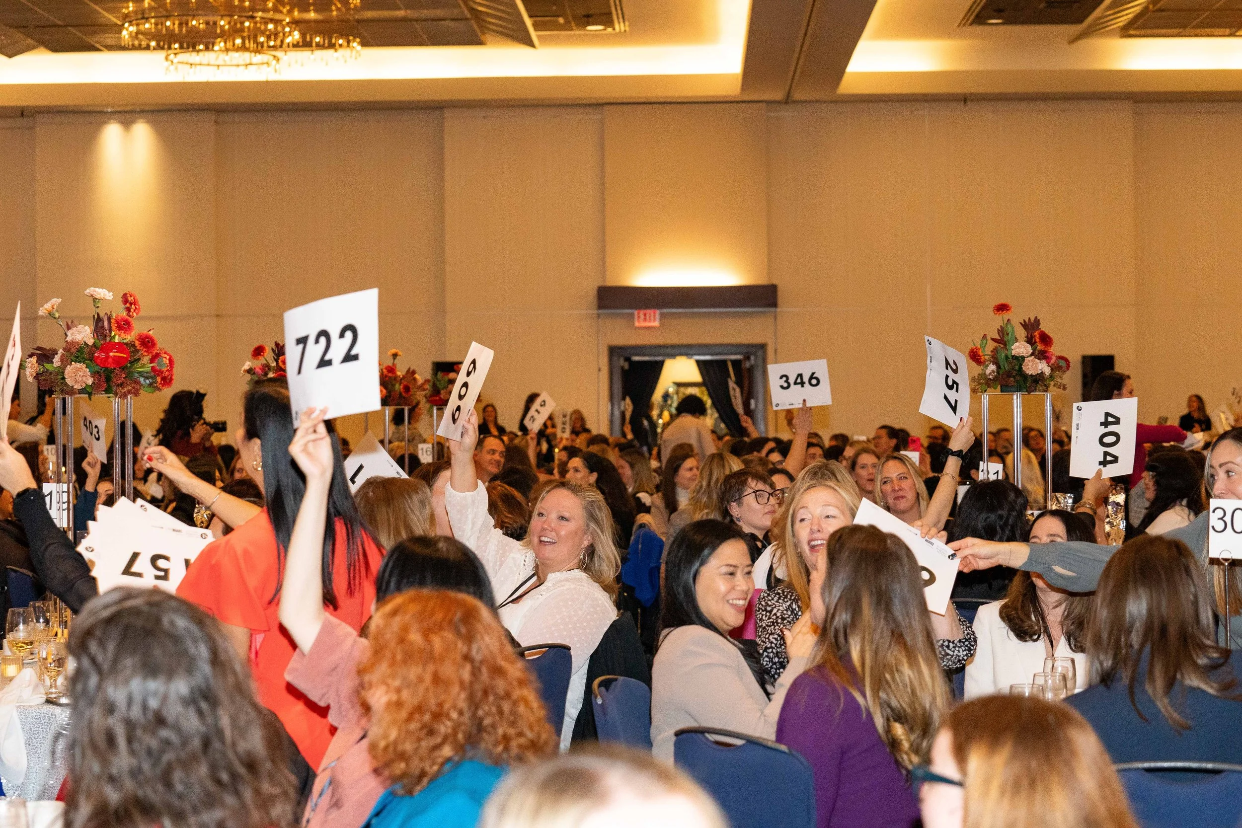 People at a large indoor event holding up numbered signs with floral centerpieces on tables.