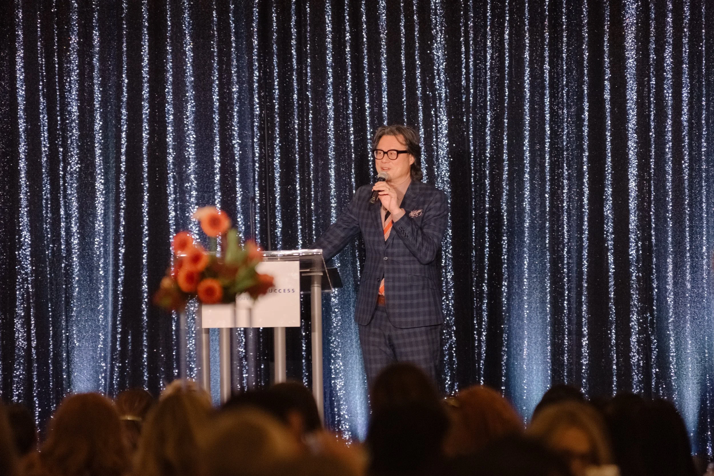 A man in a checked suit and glasses speaking at a podium with a microphone, in front of a black and silver glittery curtain, with an audience and a blurred bouquet of orange flowers in the foreground.