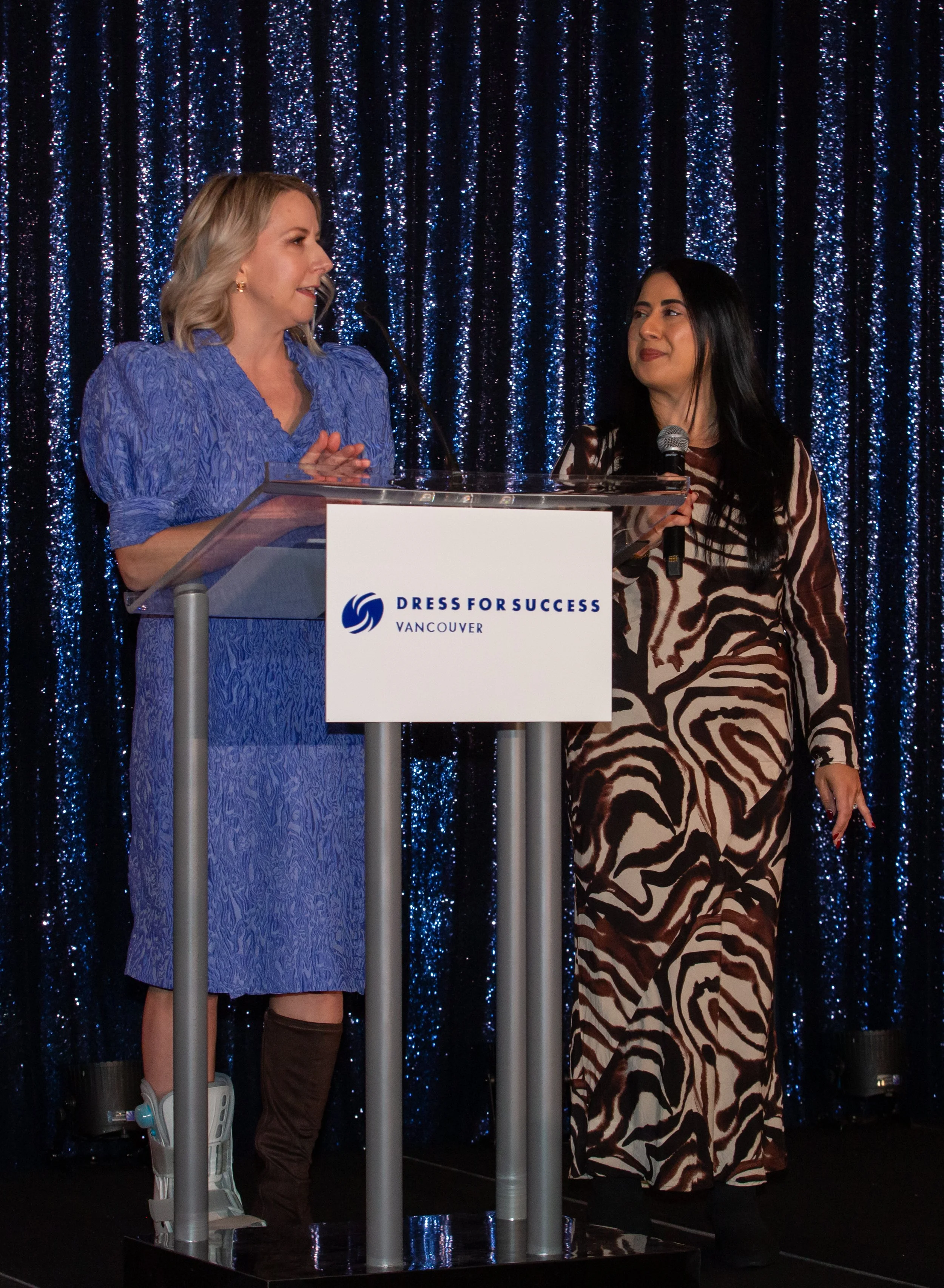 Two women speaking on a stage at a 'Dress for Success' event in Vancouver, standing behind a podium with a blue sequin curtain backdrop. One woman is in a blue dress and the other in a zebra print dress, both holding microphones.