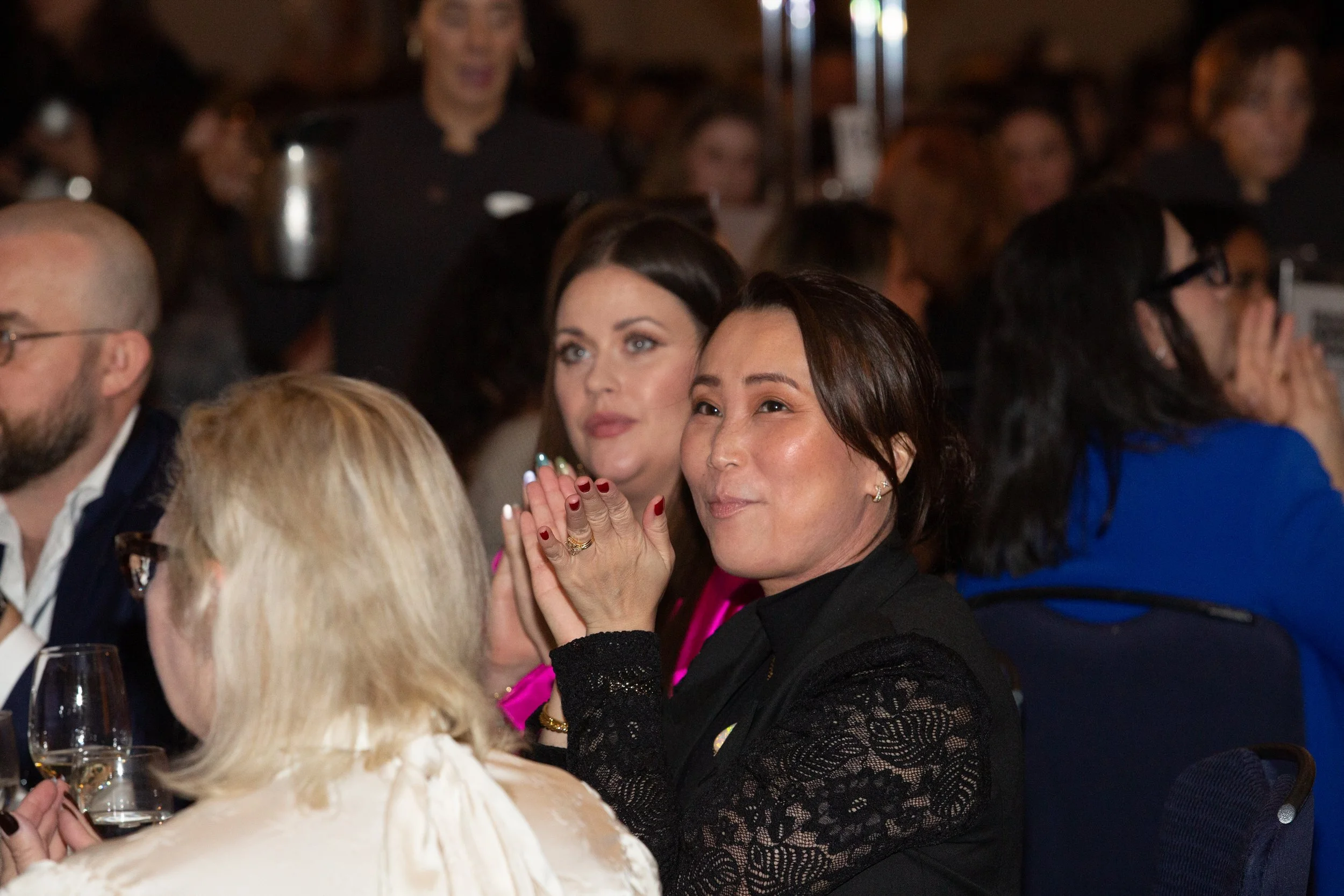 Crowd of people seated at a formal event, some clapping and watching intently, with a woman in black lace actively clapping and smiling.