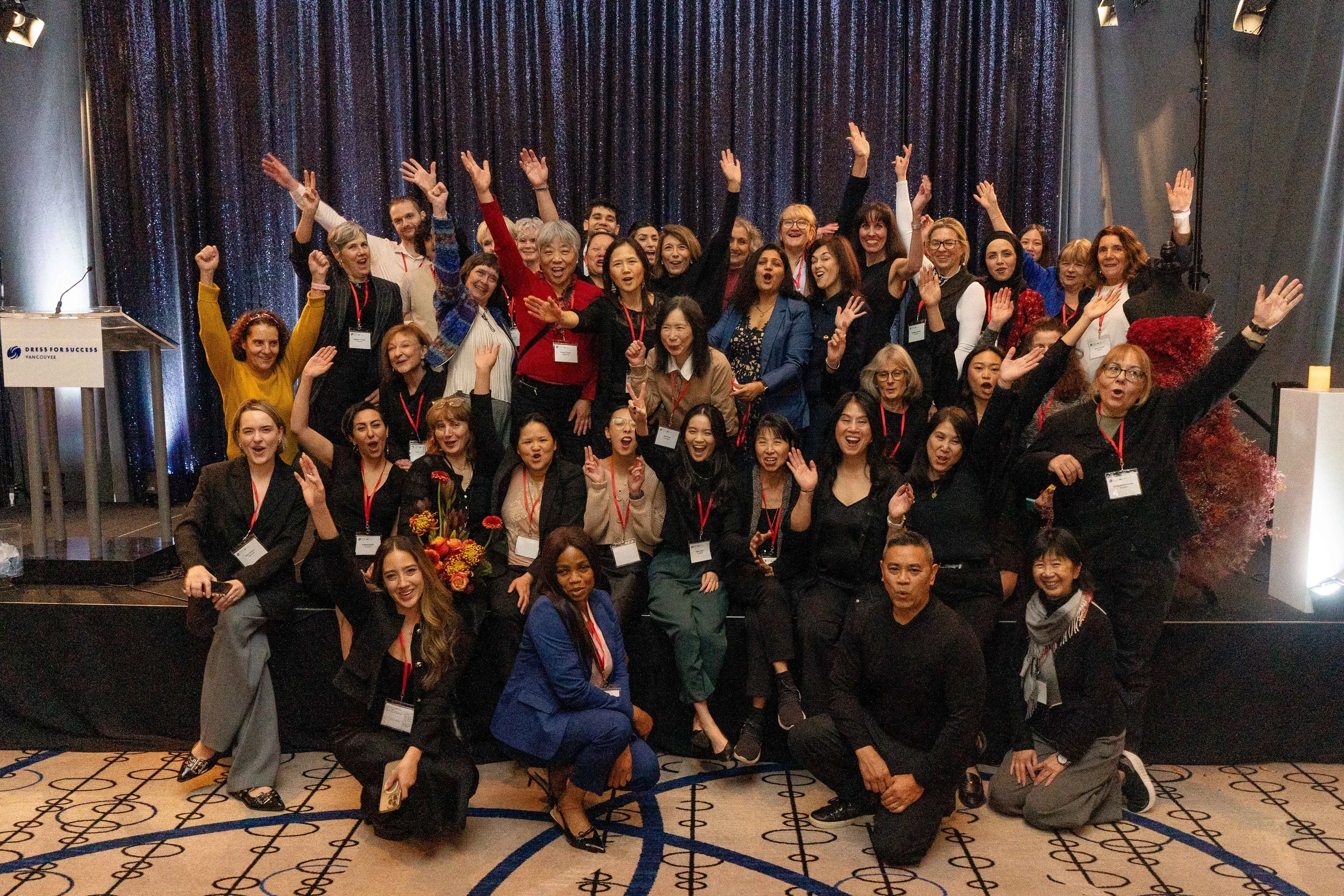 A large group of diverse women and men posing together on a stage at a conference or event. They are smiling, some with arms raised or making peace signs, in front of a dark curtain and on a patterned carpet.