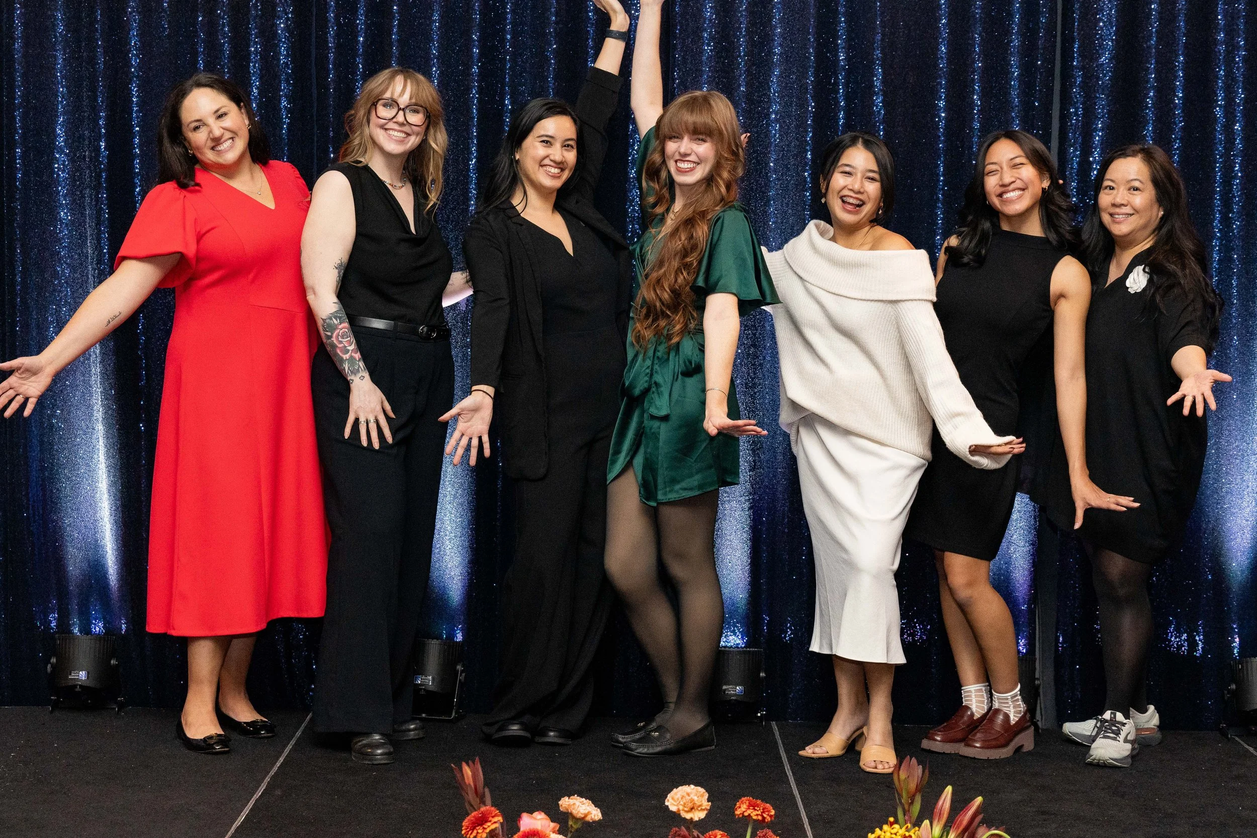 A diverse group of nine women standing on stage with blue sparkly backdrop, smiling and posing with one woman raising her arm, dressed in formal and semi-formal attire.