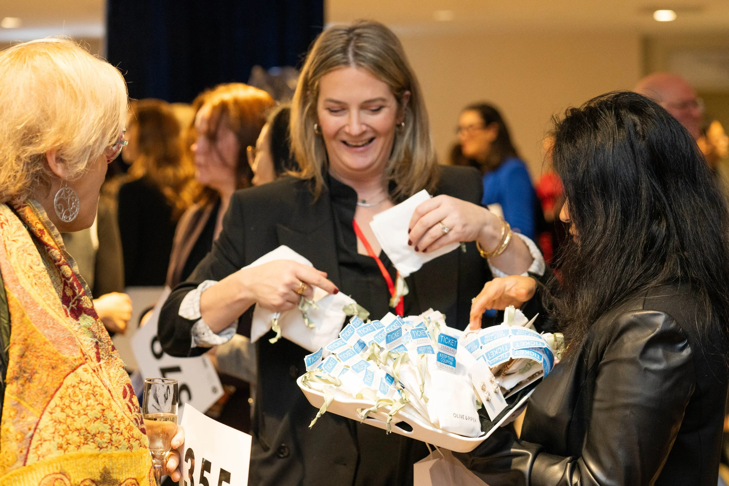 Three women at a social event exchanging small gift bags, with one holding a glass of champagne, in a crowded indoor setting.
